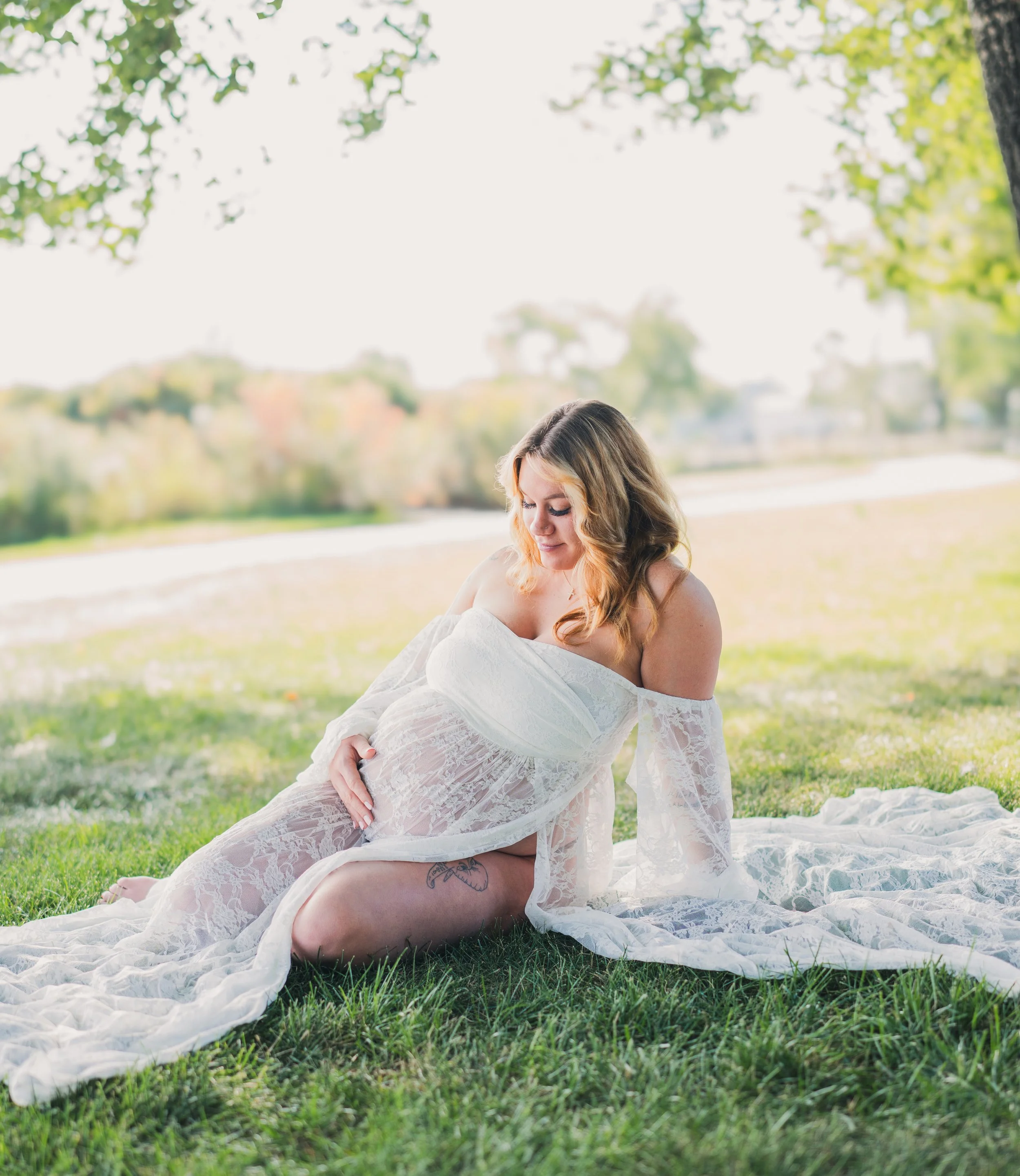 A pregnant woman sitting on a white lace blanket under a tree on a grassy field, with water and trees in the background, looking down at her belly.