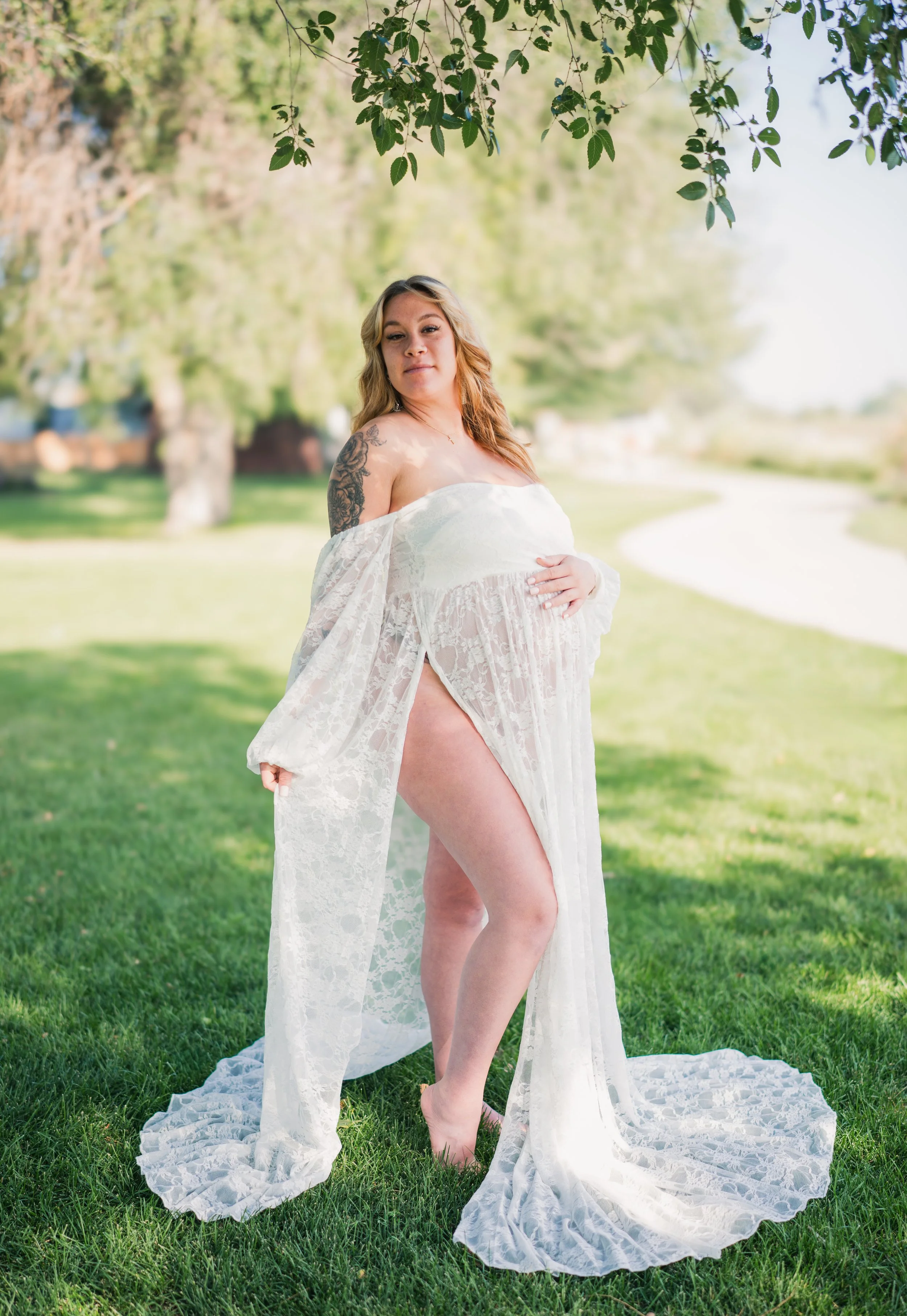 A pregnant woman stands on grass in a sheer white lace dress outdoors, with trees and a clear sky in the background.