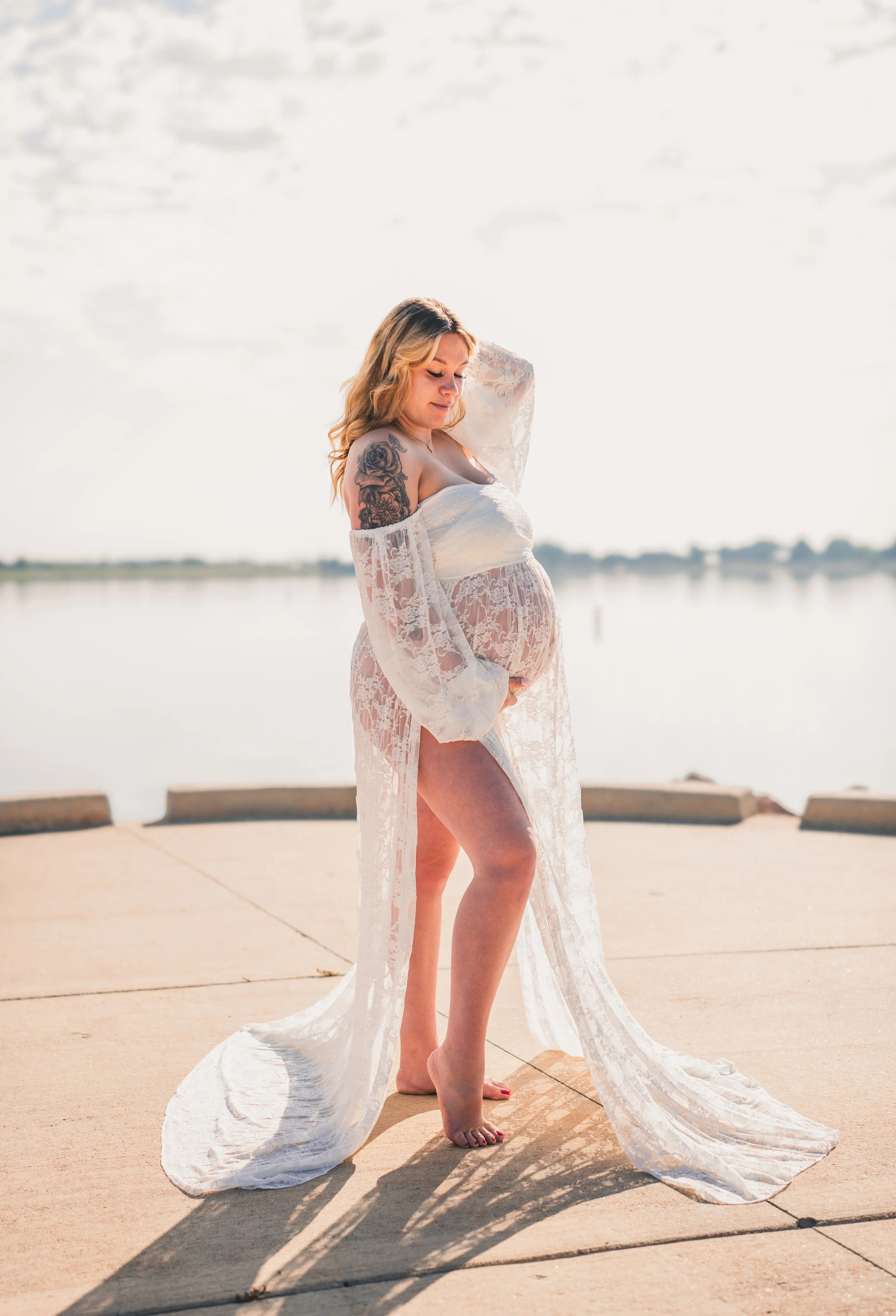 Pregnant woman outdoors on a paved surface near water, wearing white lace robe, holding her baby bump, with long wavy blonde hair and a floral tattoo on her left arm, looking down with her hand resting on her head.