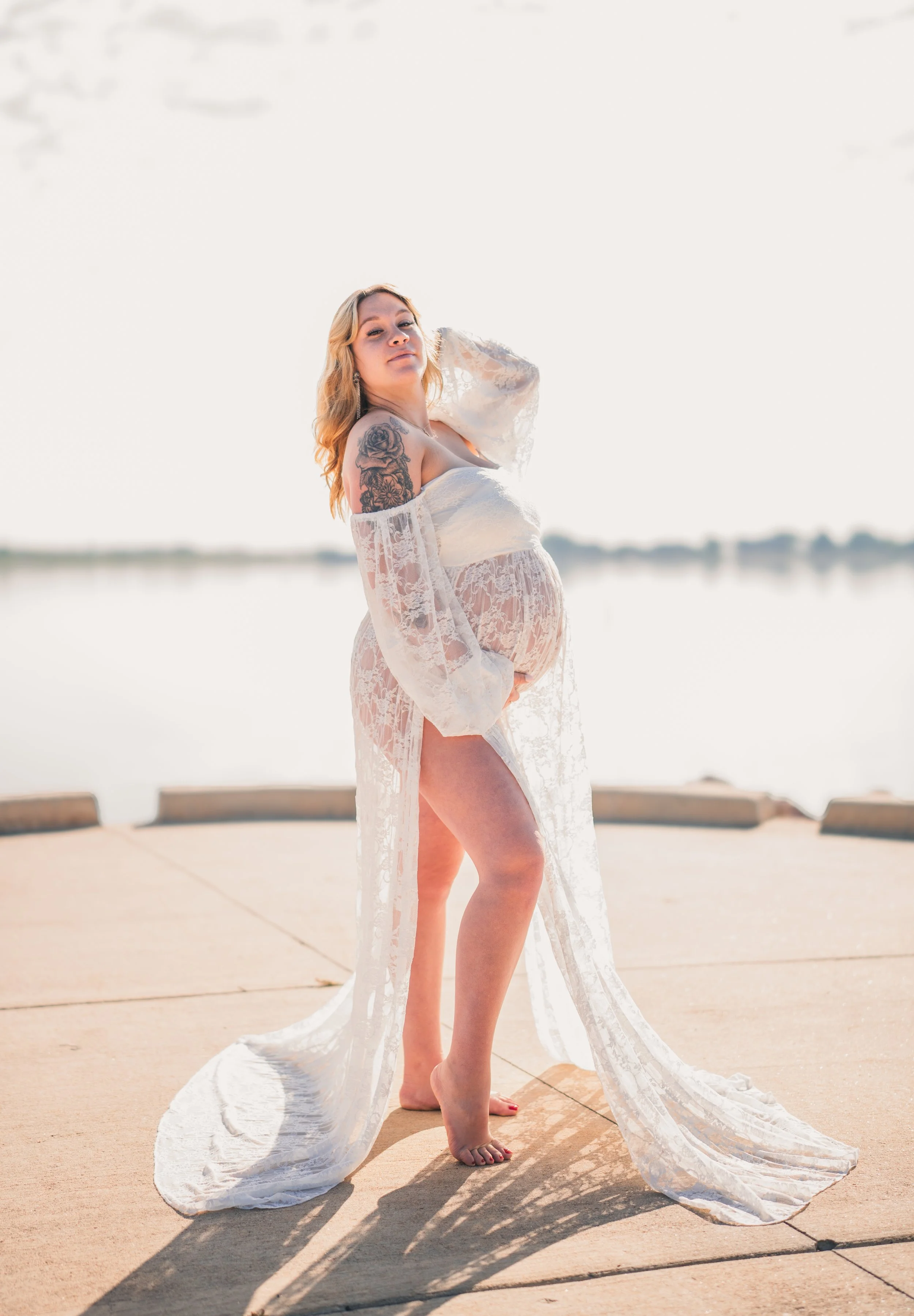 A pregnant woman in a white lace dress standing barefoot on a concrete surface outdoors near a body of water, with a clear sky and trees in the background.