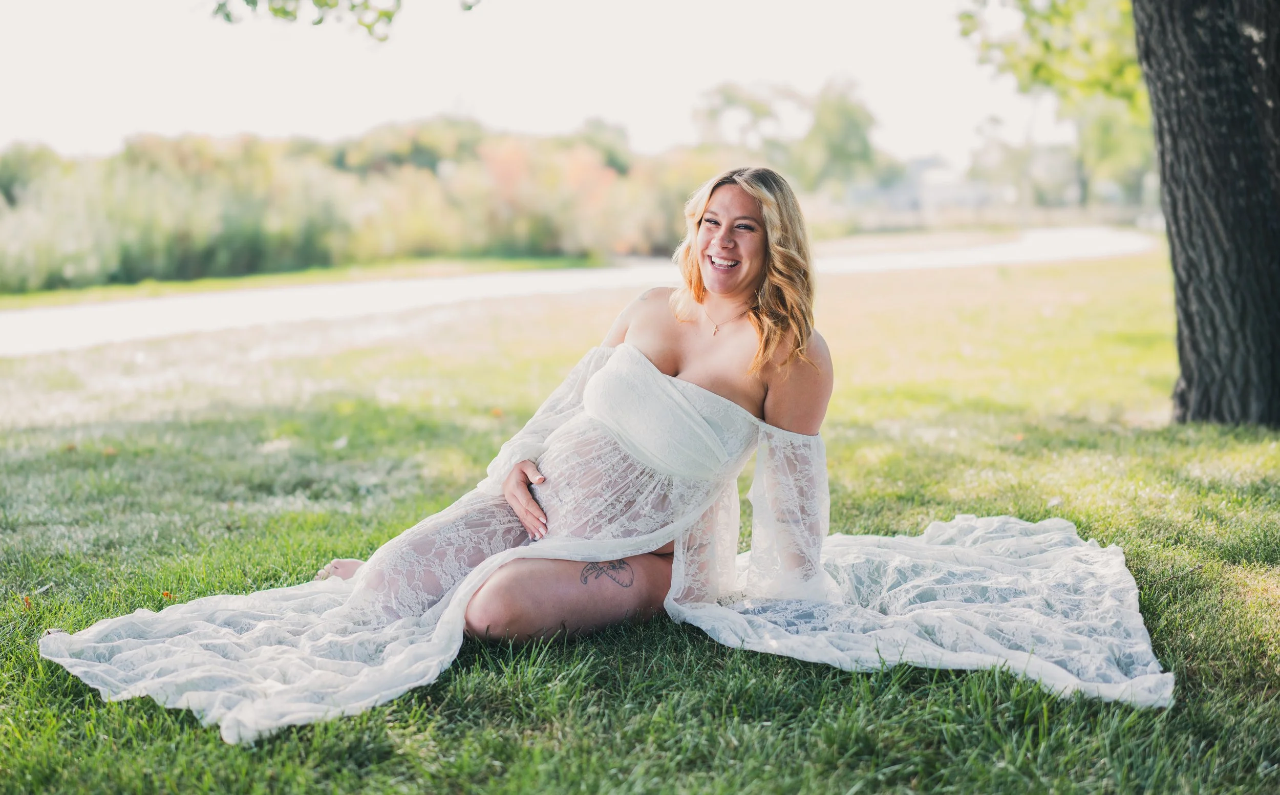 A smiling woman with blonde hair in a white lace dress sits on grass under a tree near a body of water in a park on a sunny day.