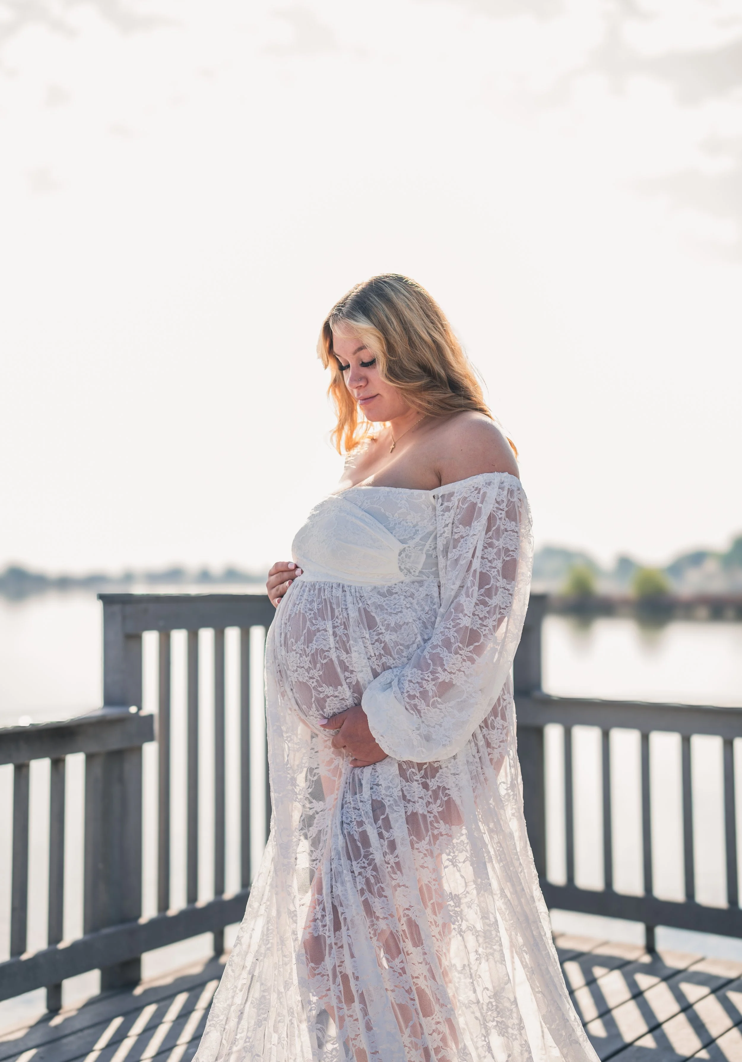 Pregnant woman in white lace dress standing on a wooden dock near water during sunset.