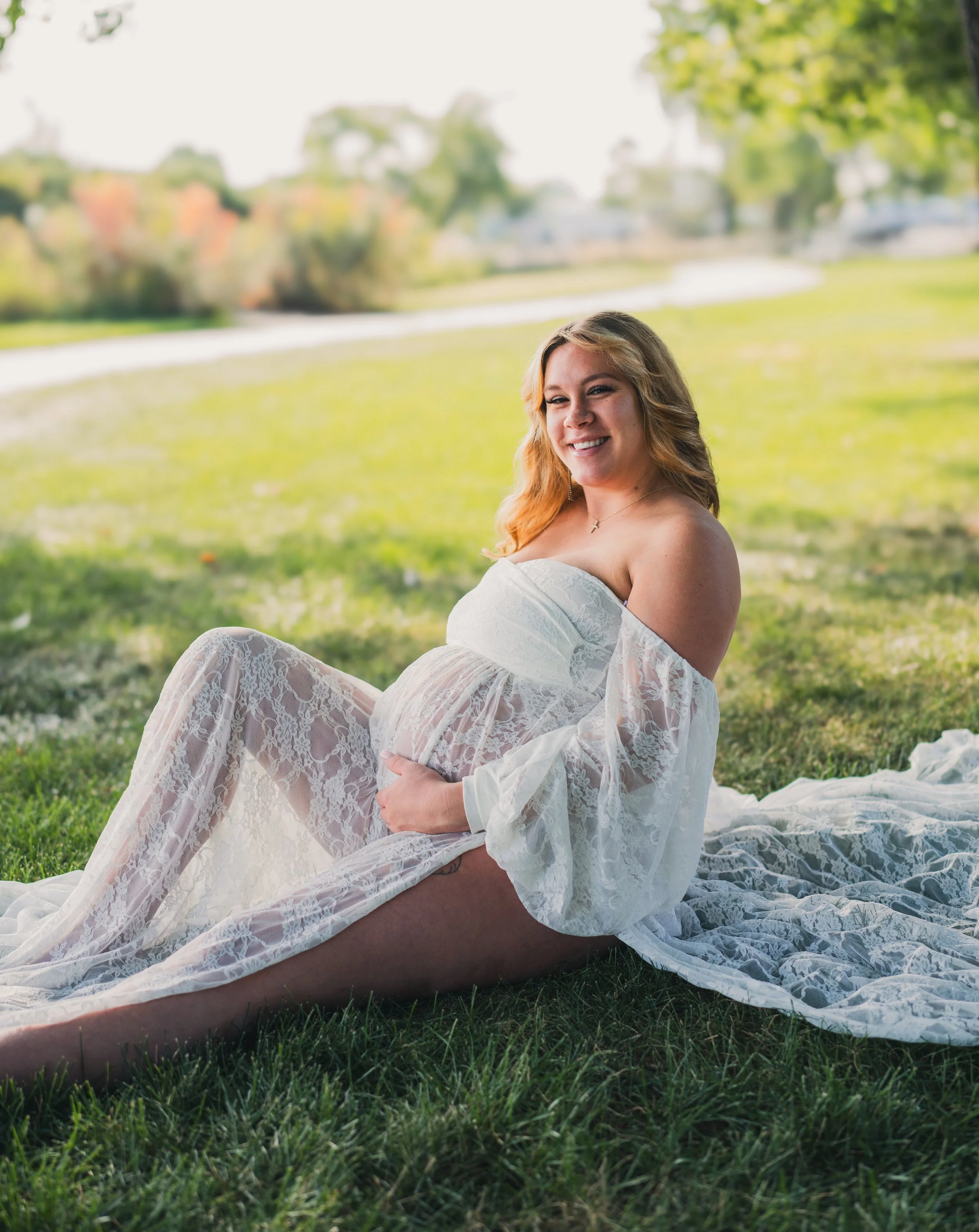 A pregnant woman sitting on the grass in a park, wearing a white lace off-the-shoulder dress, smiling and looking at the camera with a pond and trees in the background.