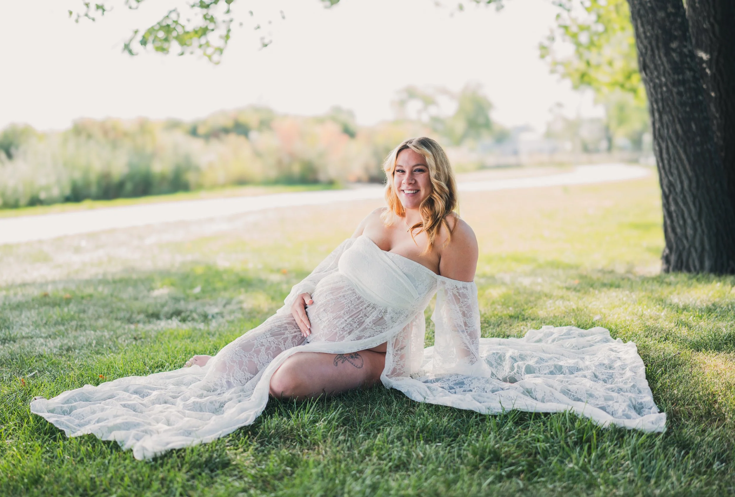 A pregnant woman with red hair, wearing a white sheer lace dress, is sitting on the grass under a large tree in a park, smiling and looking off to the side with a happy expression.