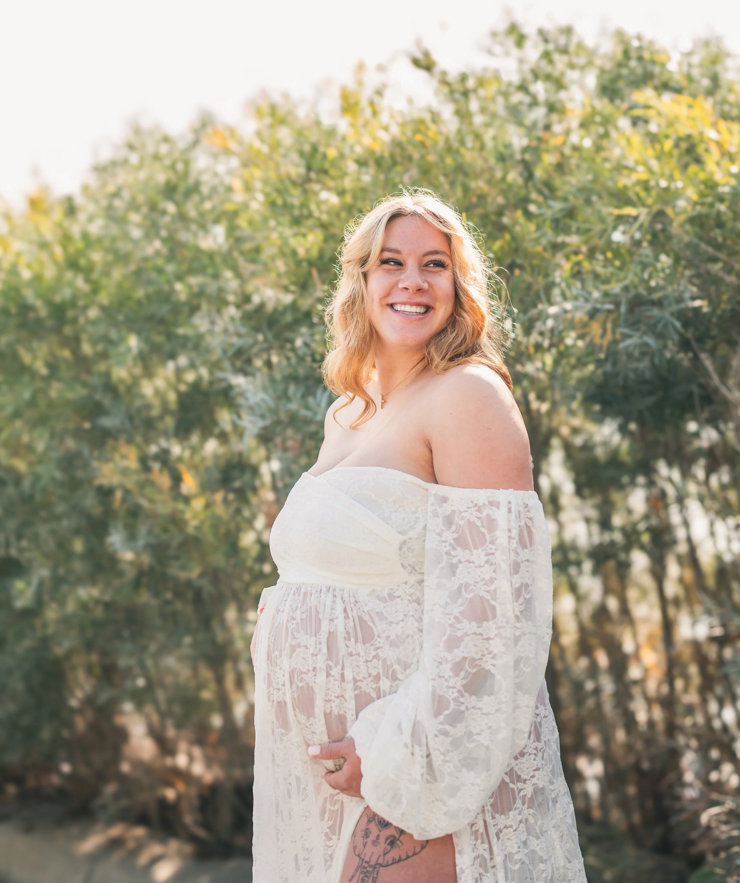 Young woman with wavy blonde hair smiling outdoors, wearing a white lace off-the-shoulder dress, standing against a background of trees and sunlight.