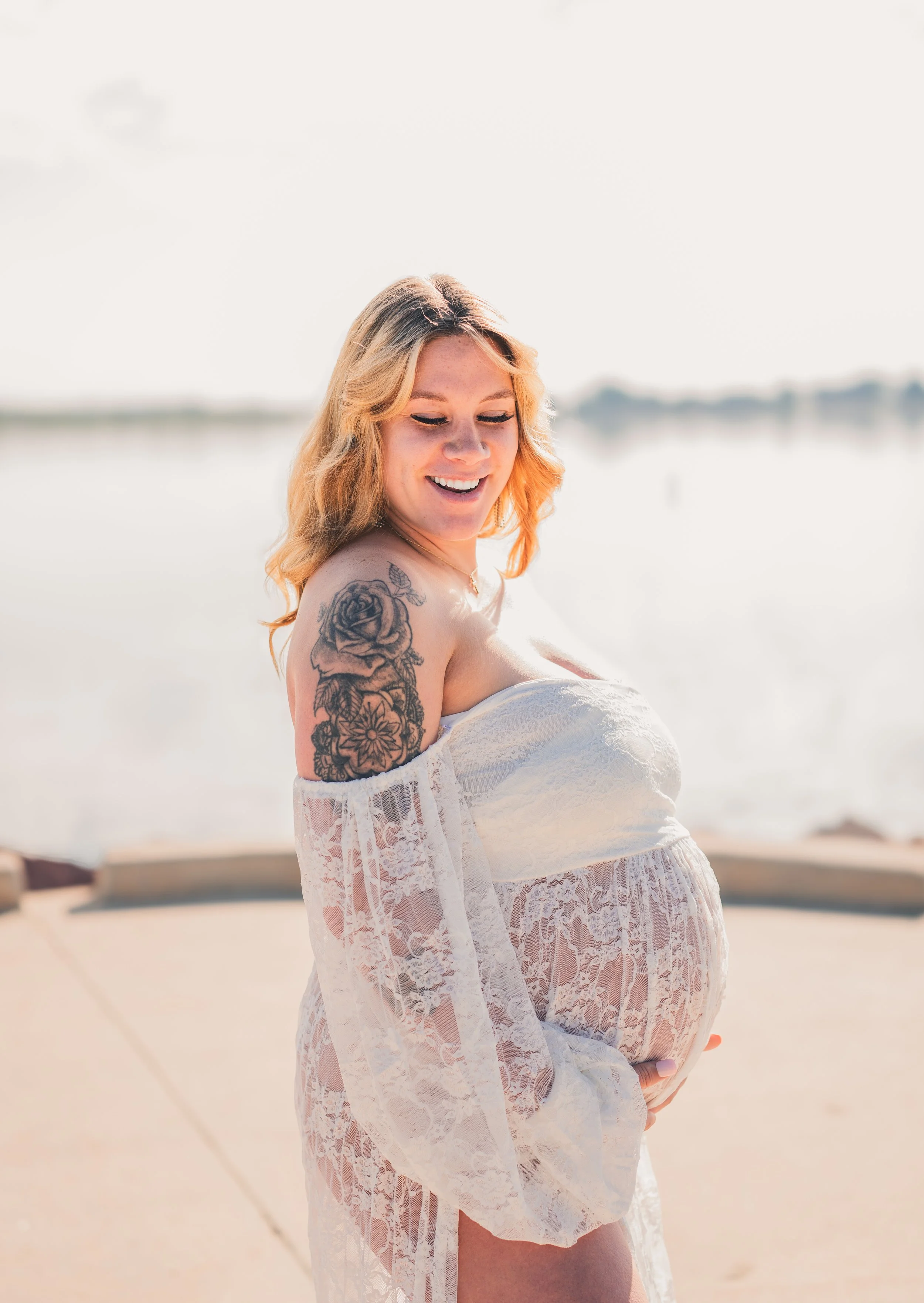 A pregnant woman with blonde hair and a floral tattoo on her arm, wearing a white lace off-shoulder dress, smiling and holding her belly near a body of water on a sunny day.