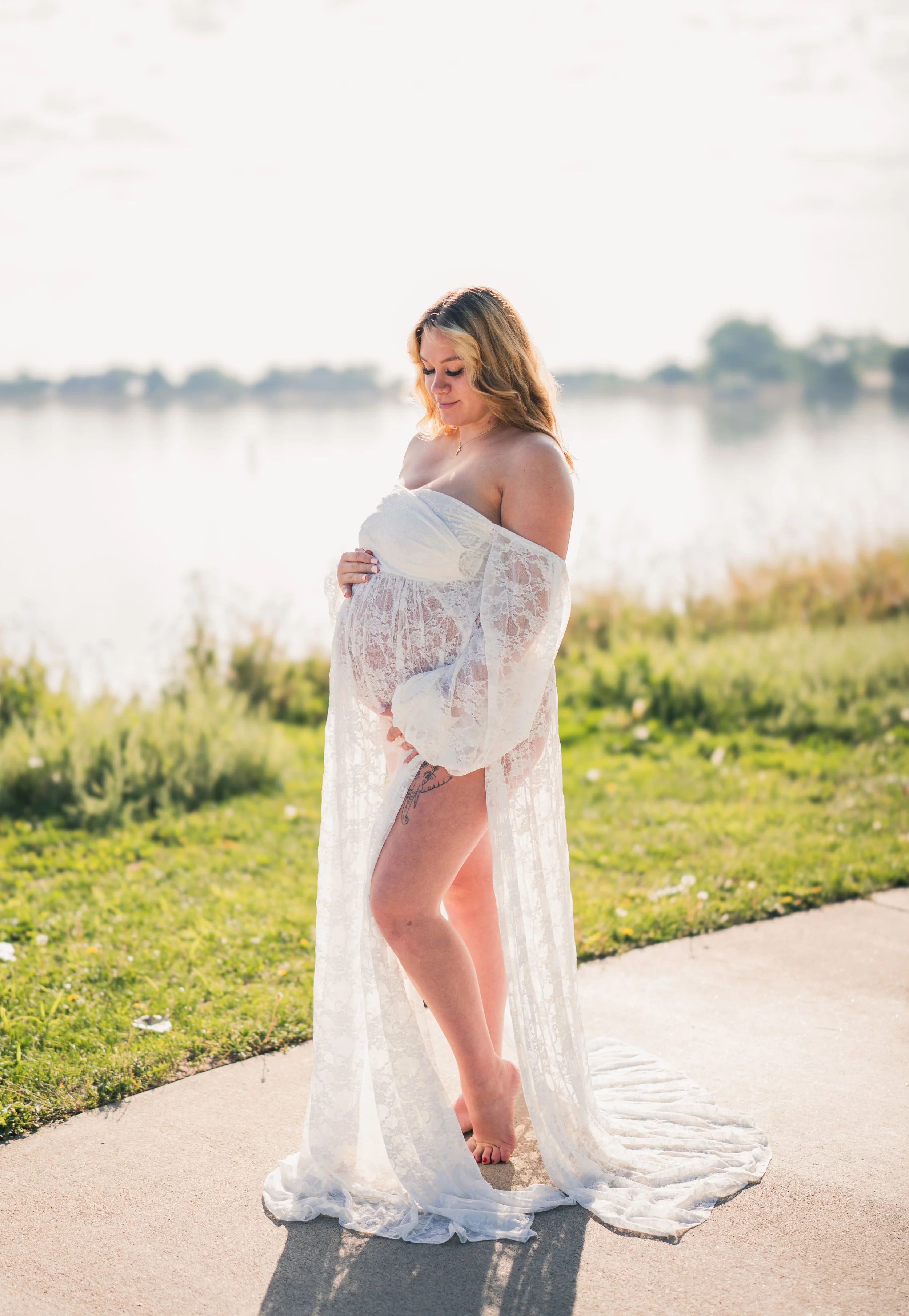 Pregnant woman in a white lace dress standing outdoors near a body of water, looking down, holding her belly, with the sunlight shining behind her.