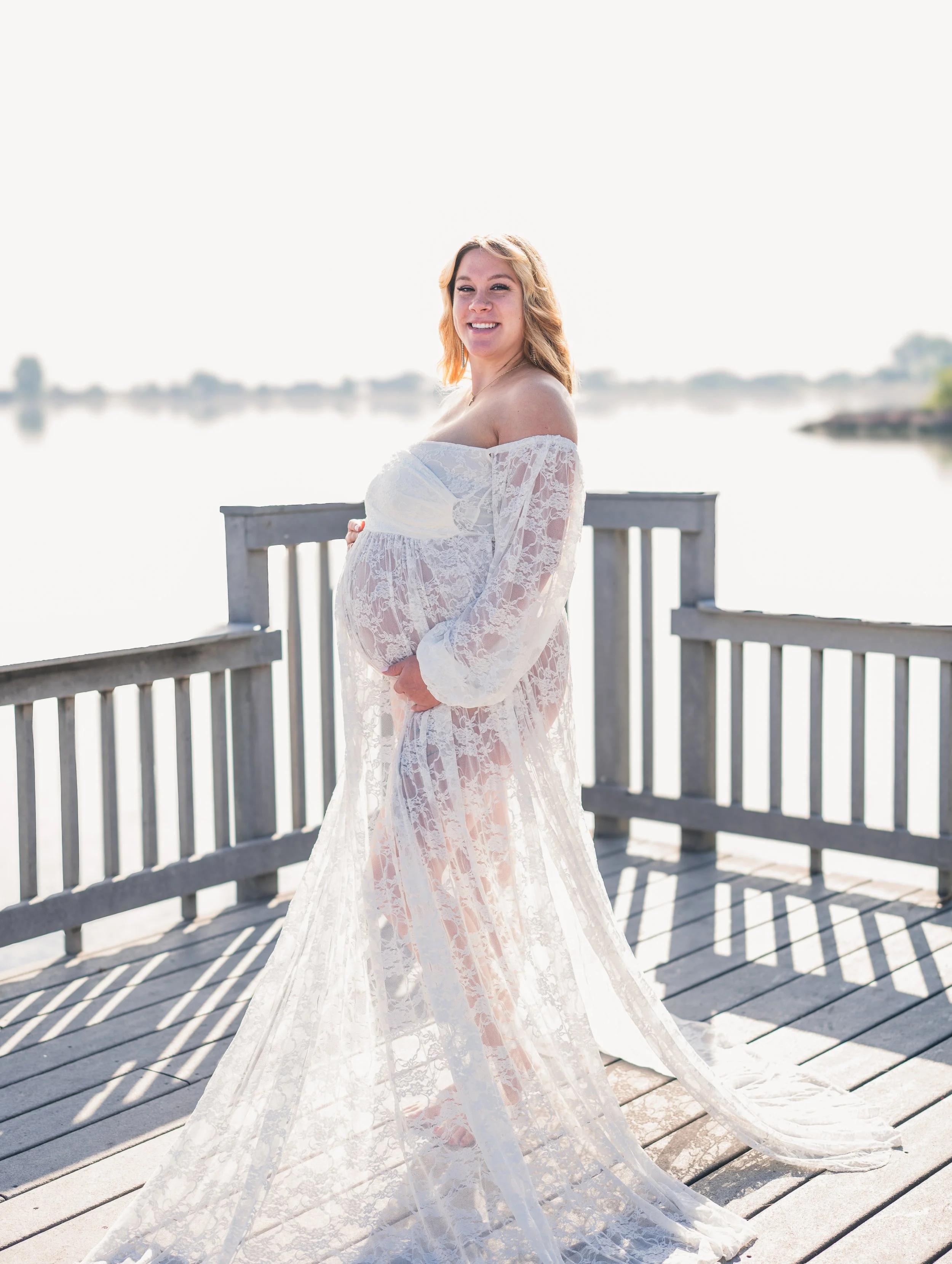 Pregnant woman in a white lace dress standing on a wooden dock by water, smiling.