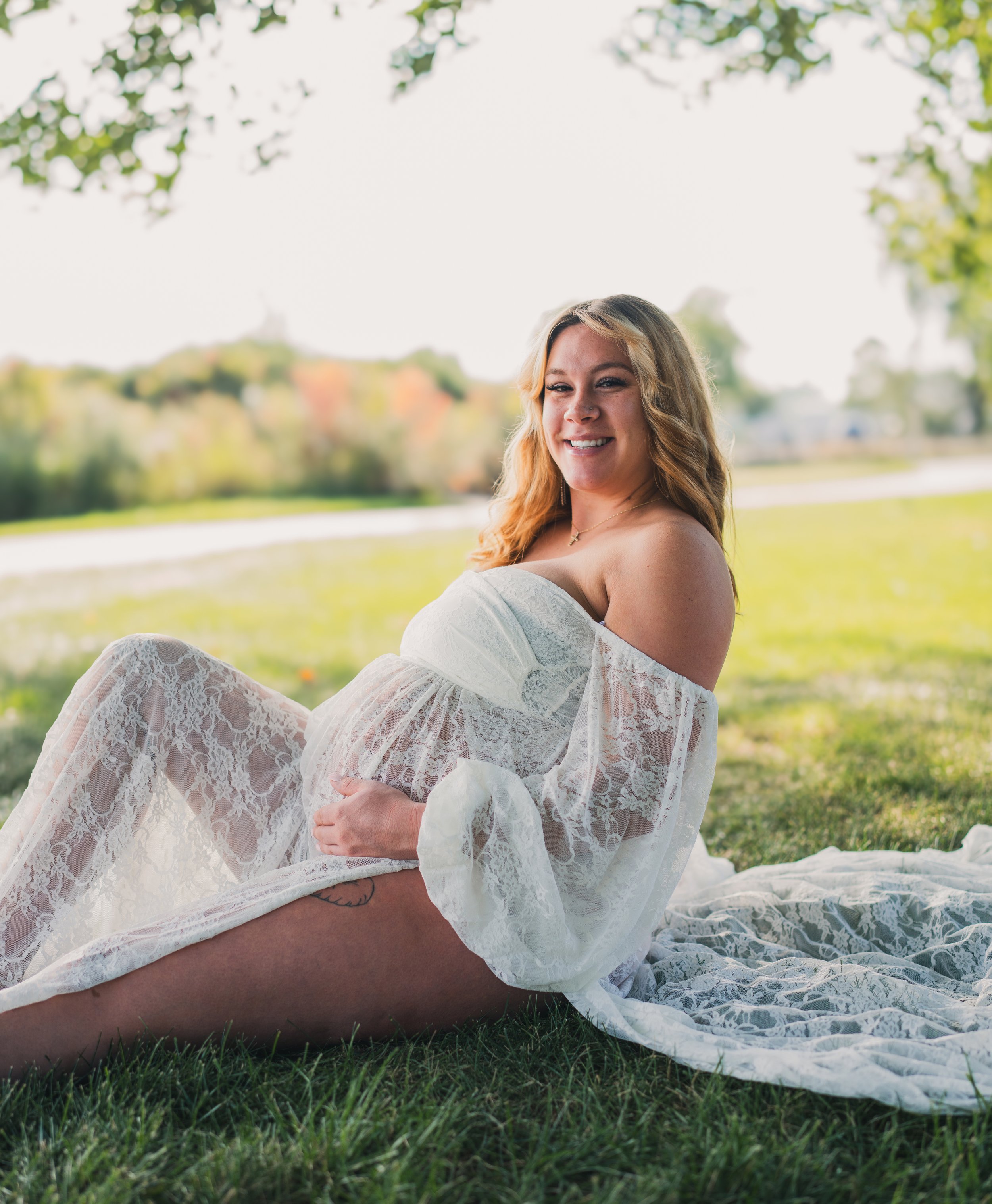 Pregnant woman in a white lace dress smiling while sitting on grass outdoors near a body of water.