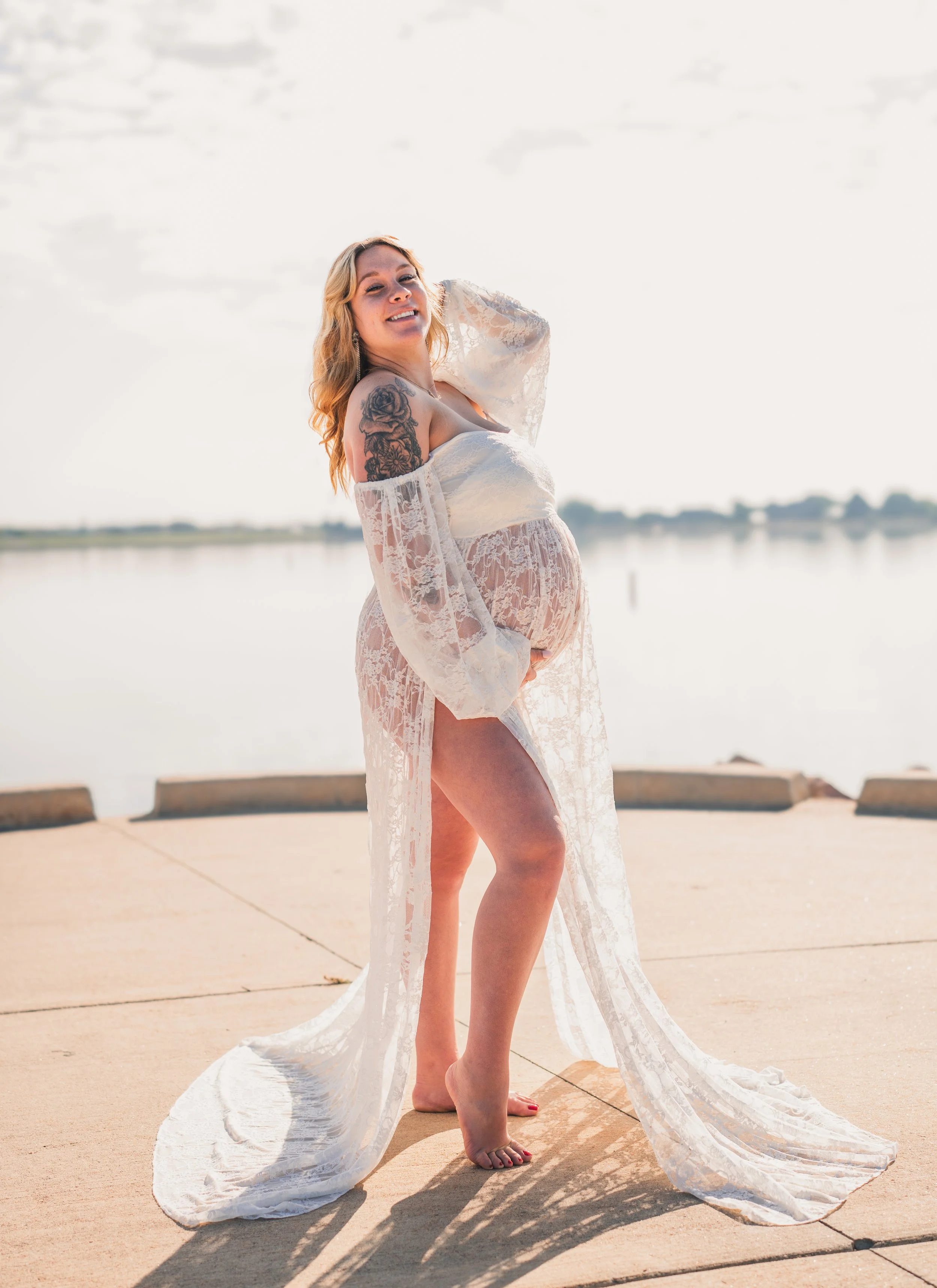 A pregnant woman in a white lace dress standing barefoot on a concrete surface near a body of water, smiling at the camera with her left hand on her belly and her right hand raised to her hair, during daytime with a cloudy sky.