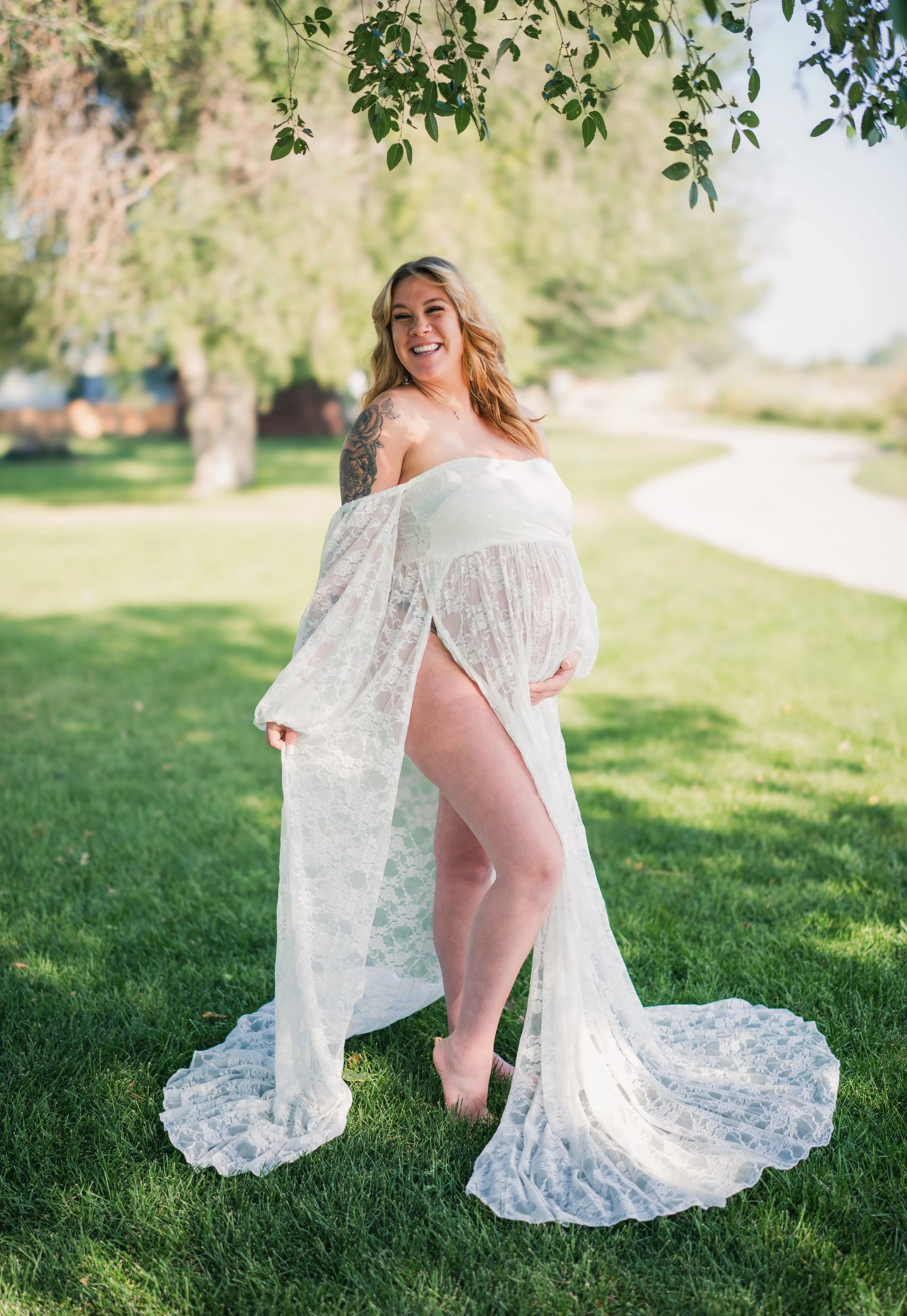 A pregnant woman standing on grass outdoors, smiling, wearing a sheer white lace dress, with trees and a pathway in the background.
