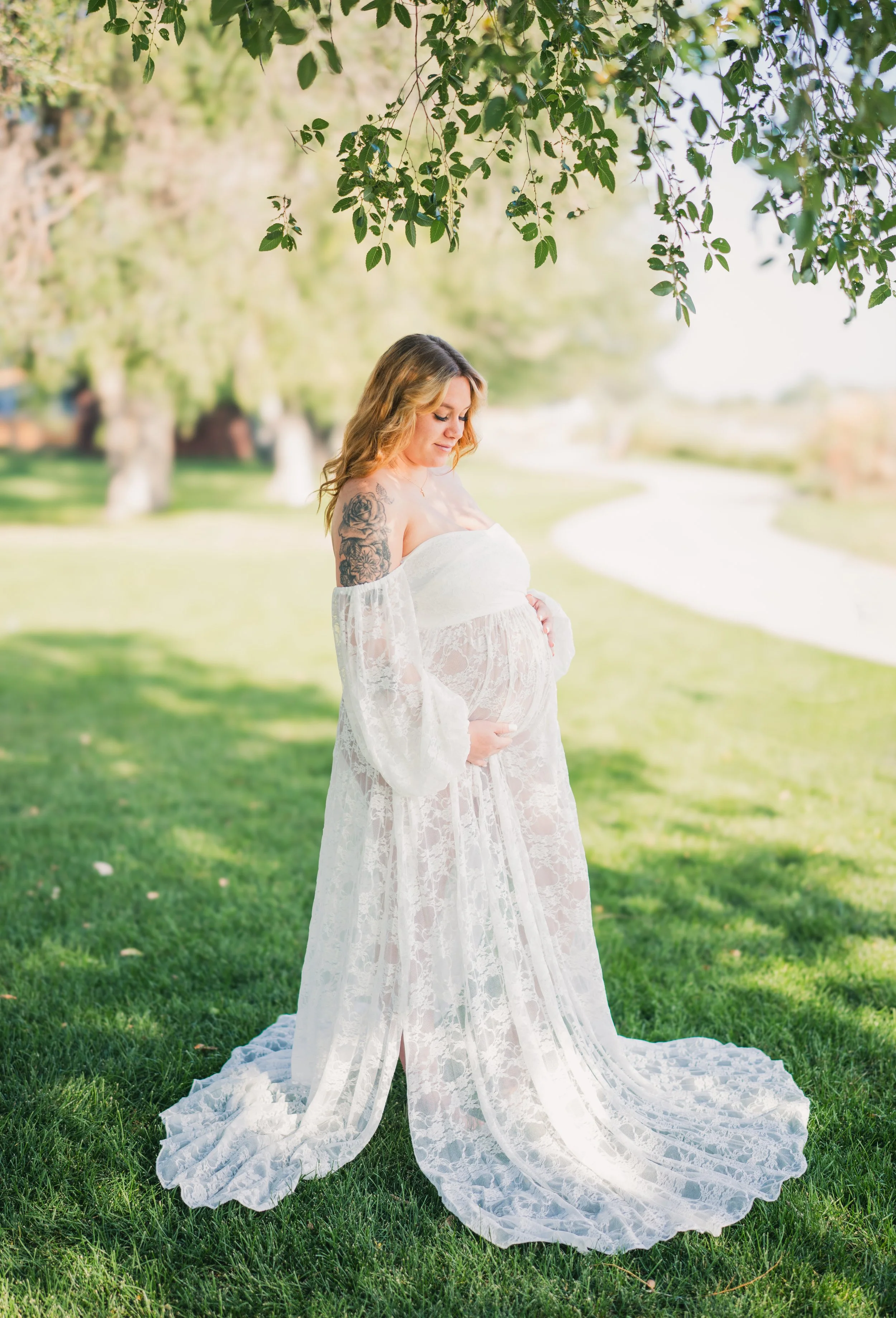 A pregnant woman in a white lace off-the-shoulder dress standing outdoors on a grassy area under a tree, with trees and a winding path in the background.