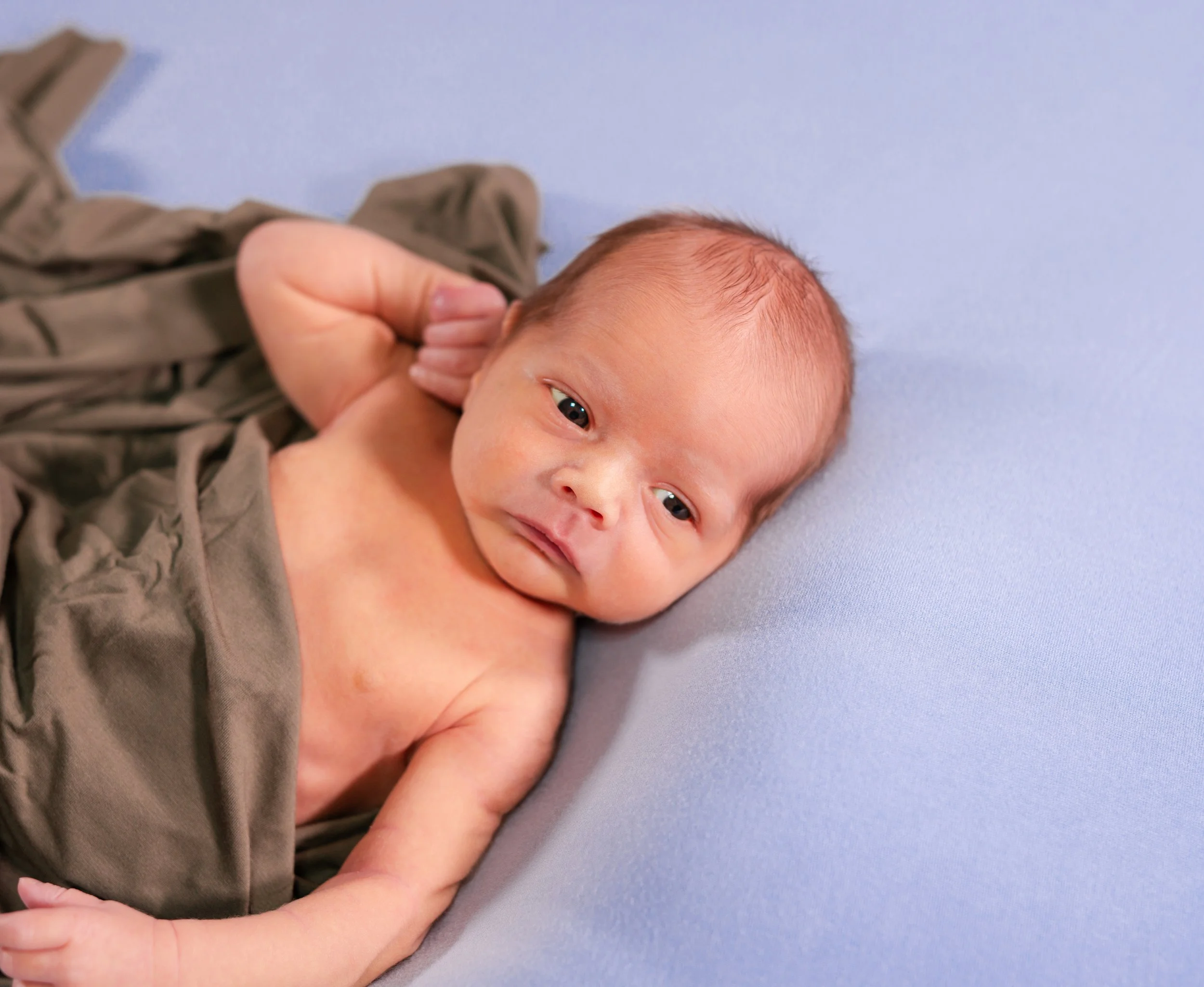 A newborn baby lying on a light blue surface, partially covered with a brown cloth, gazing at the camera with wide eyes.