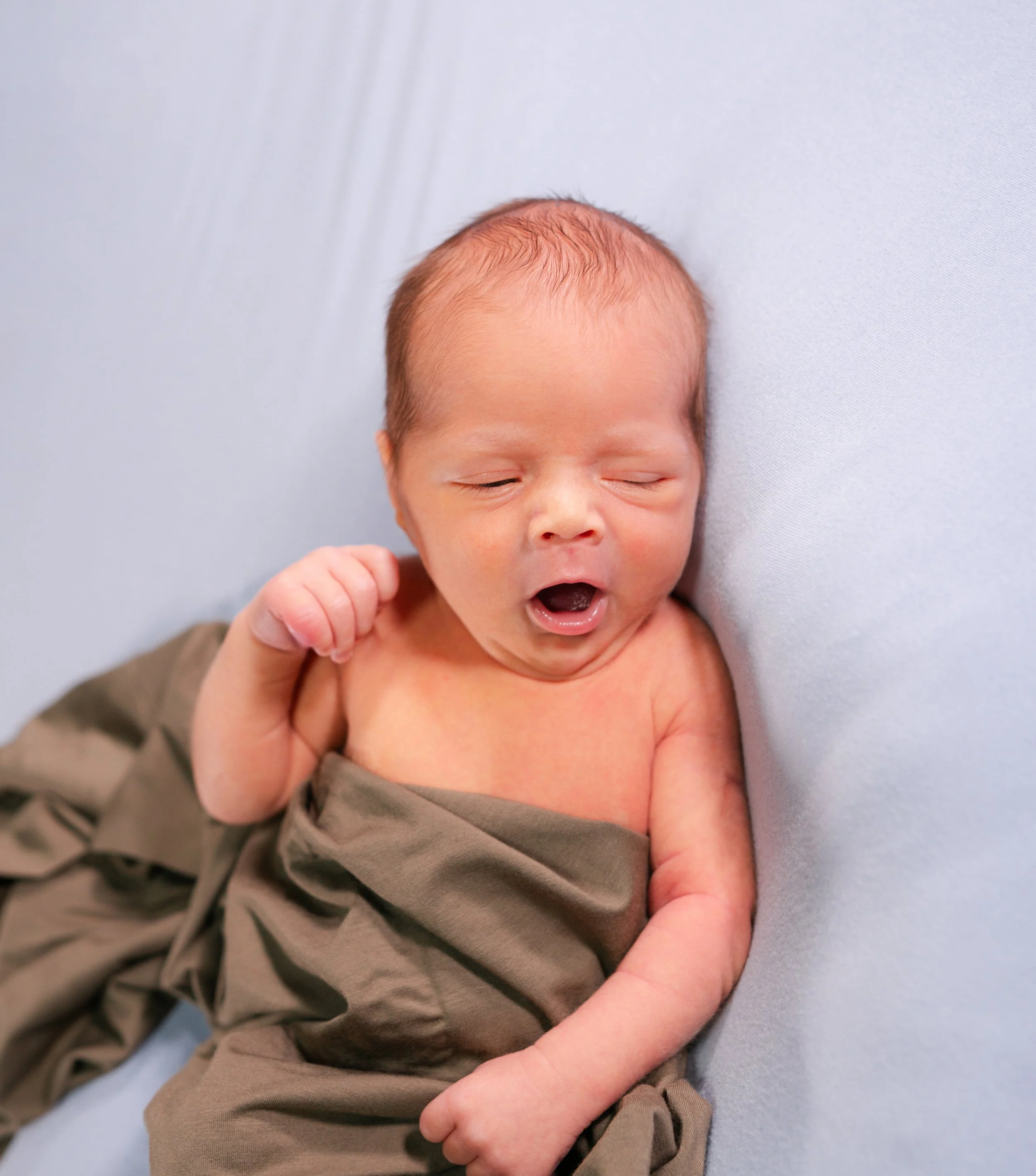 Newborn baby yawning while lying on a soft surface, wrapped in a brown cloth.