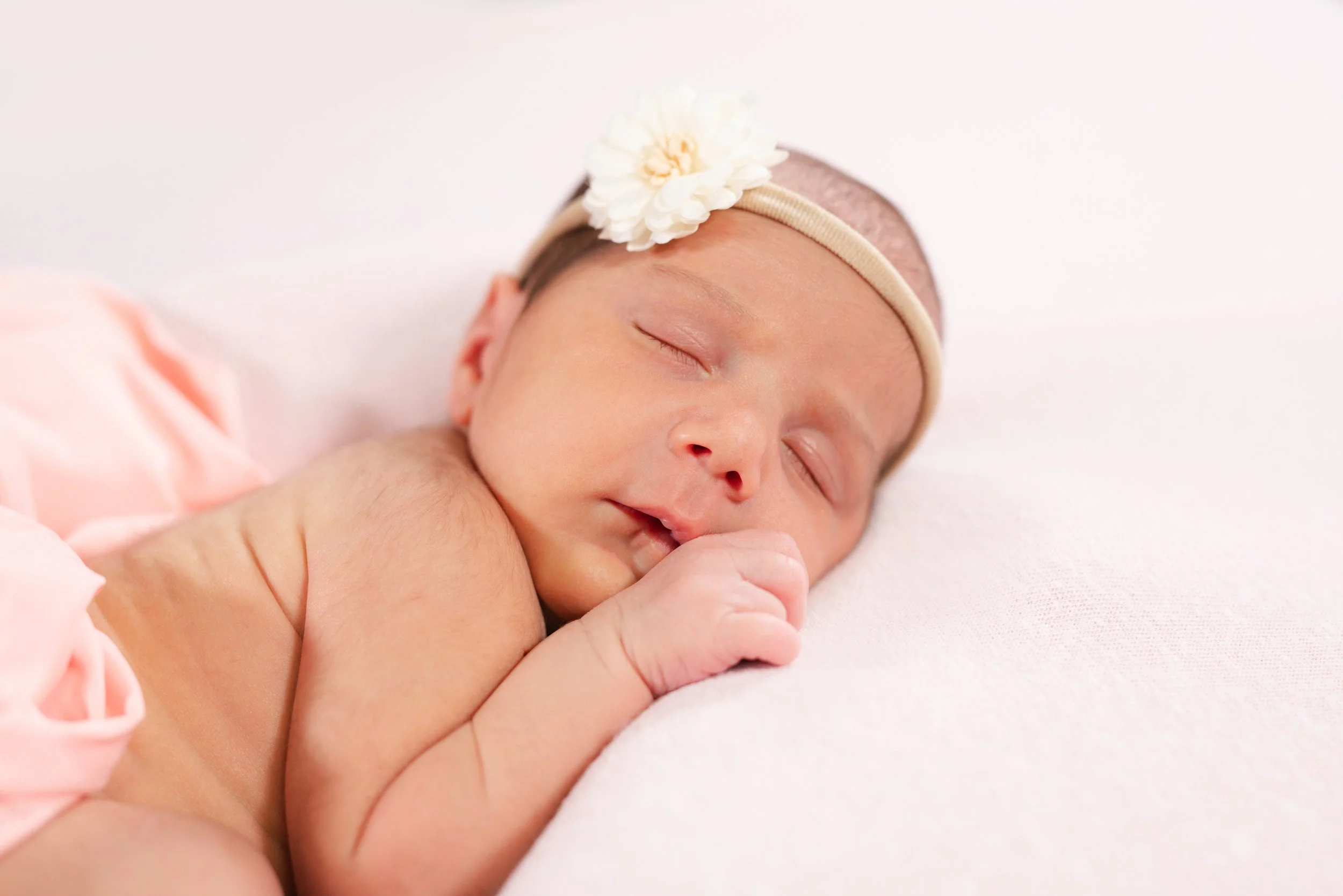 A sleeping newborn baby girl with a flower headband lying on a white surface.