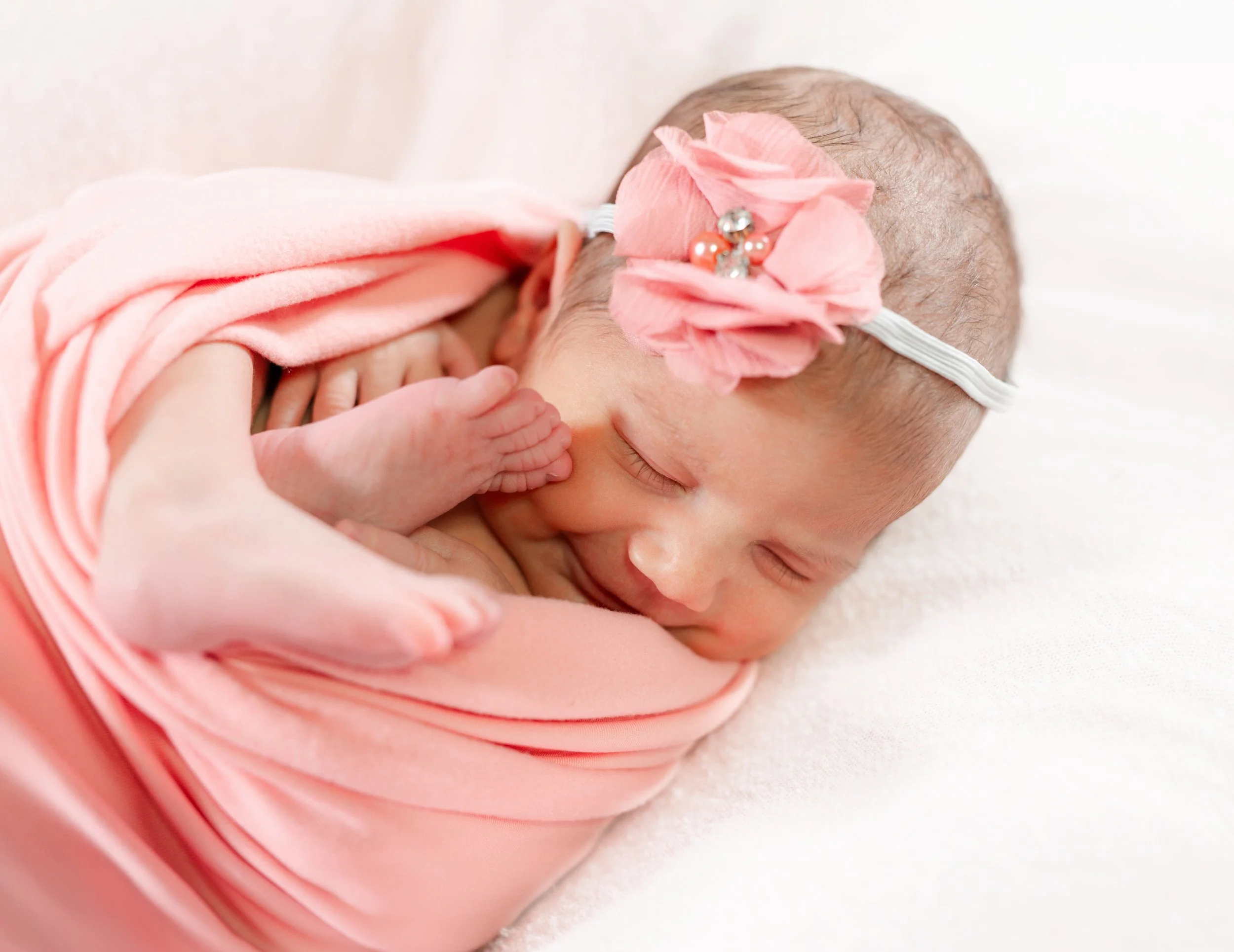 A newborn baby girl peacefully sleeping, wrapped in pink fabric, wearing a pink headband with a flower and rhinestone accents.
