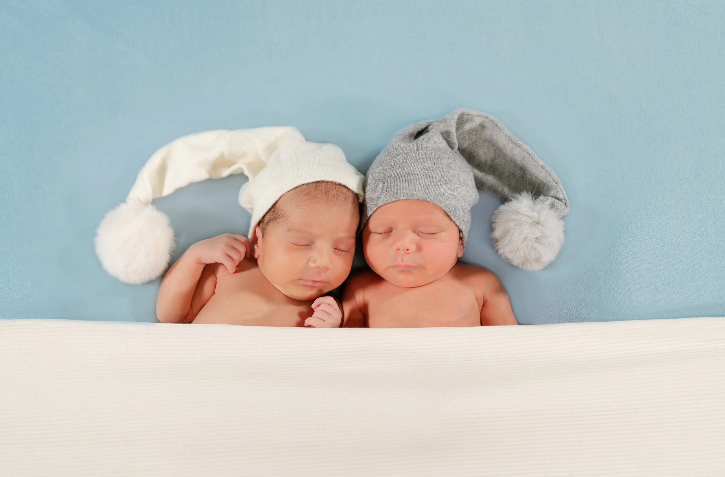 Two sleeping babies lying side by side, each wearing a knit hat with a pom-pom, on a light blue surface.