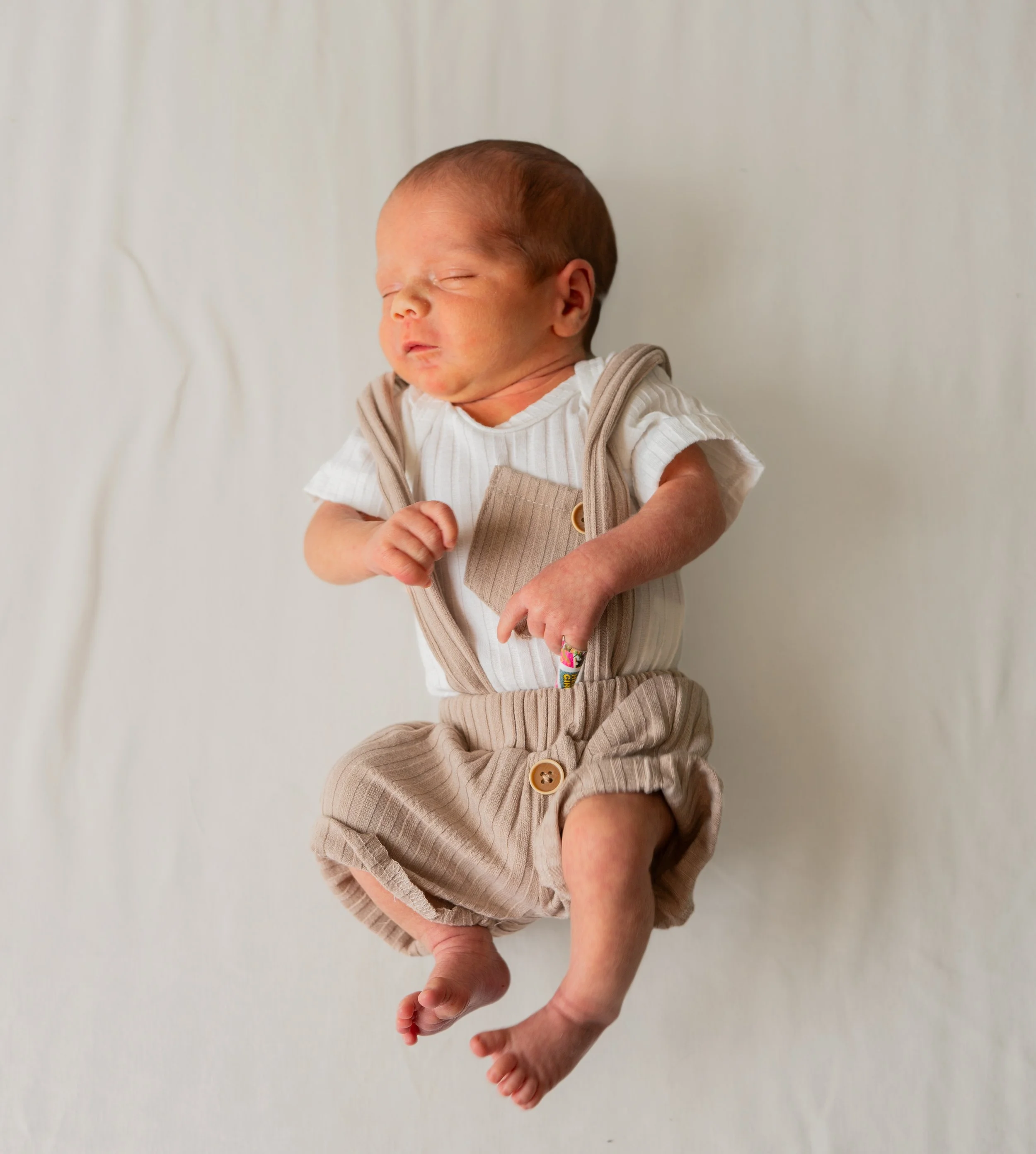 A sleeping newborn baby dressed in beige and white clothing, lying on a soft white blanket.