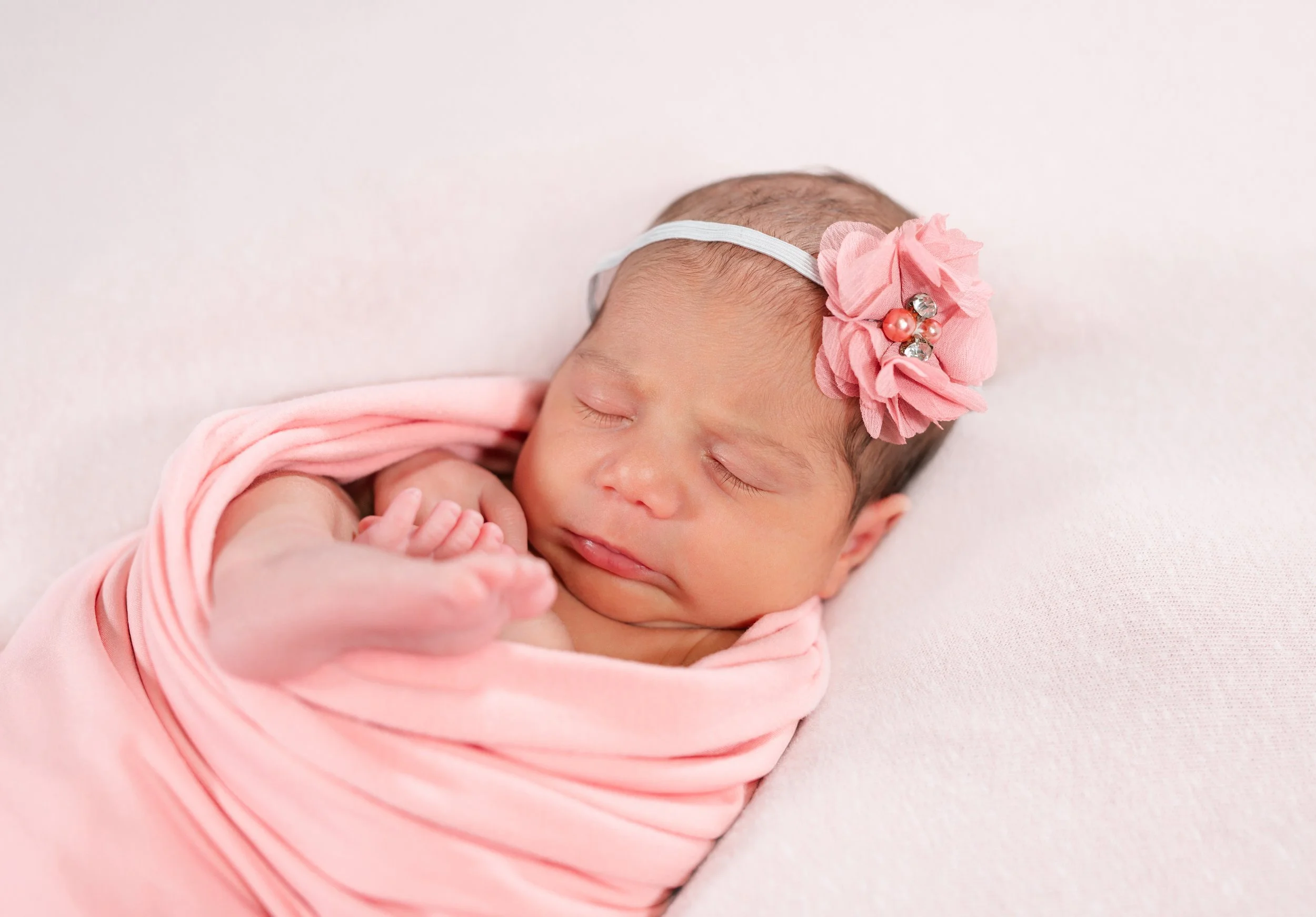 A newborn baby girl sleeping, wrapped in a pink cloth, wearing a pink headband with a fabric flower and decorative beads.