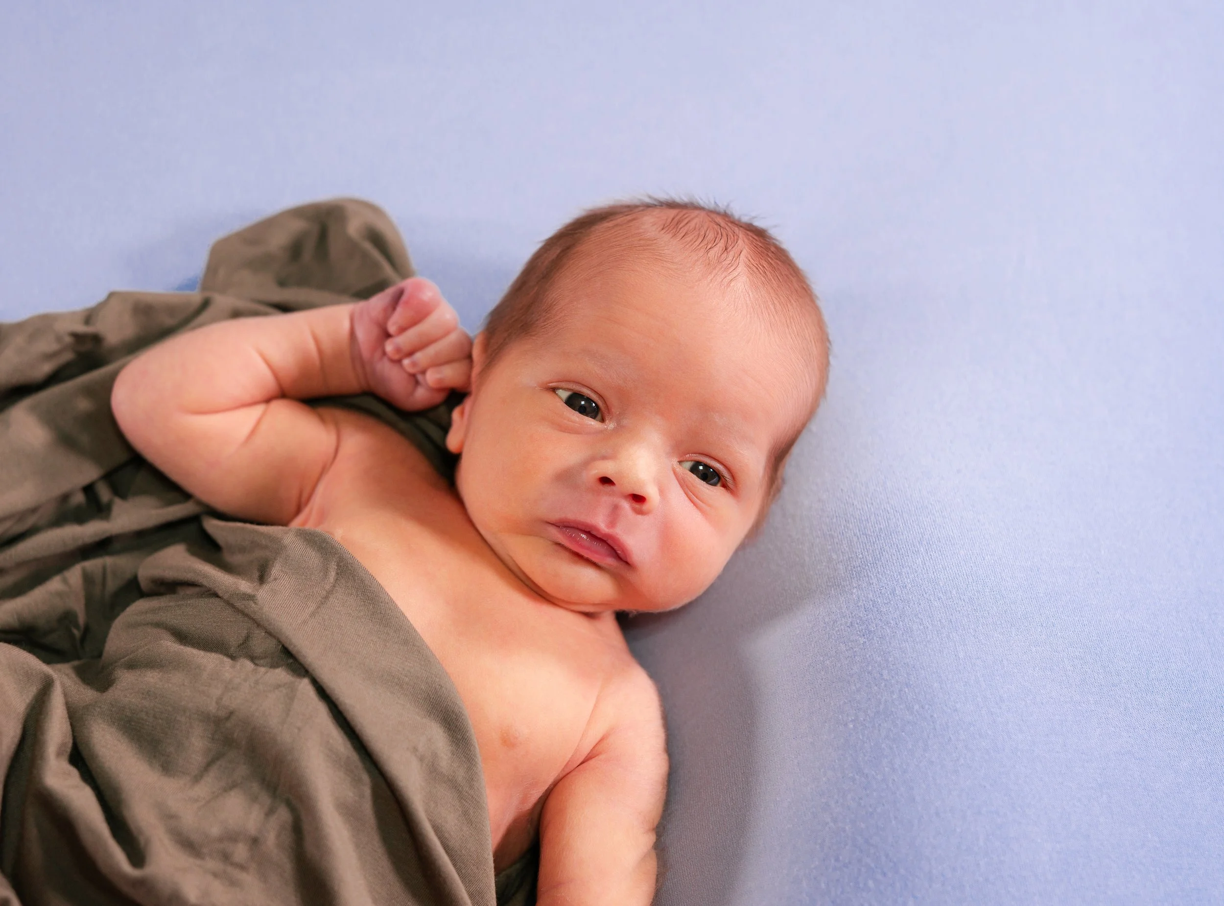 A newborn baby with blue eyes and light skin lying on a light blue surface, partially covered with a brown cloth.