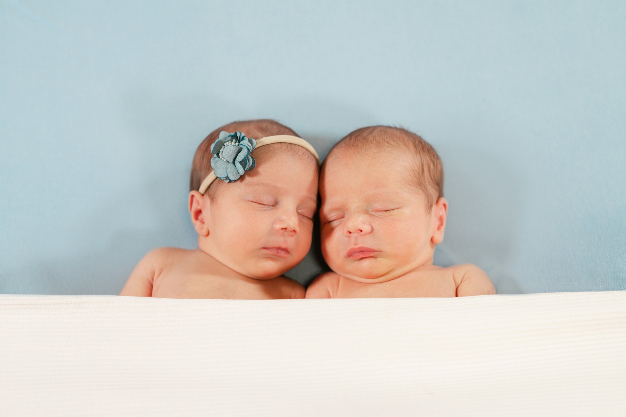 Two sleeping babies lying close together with heads touching, one wearing a headband with a flower, against a light blue background.
