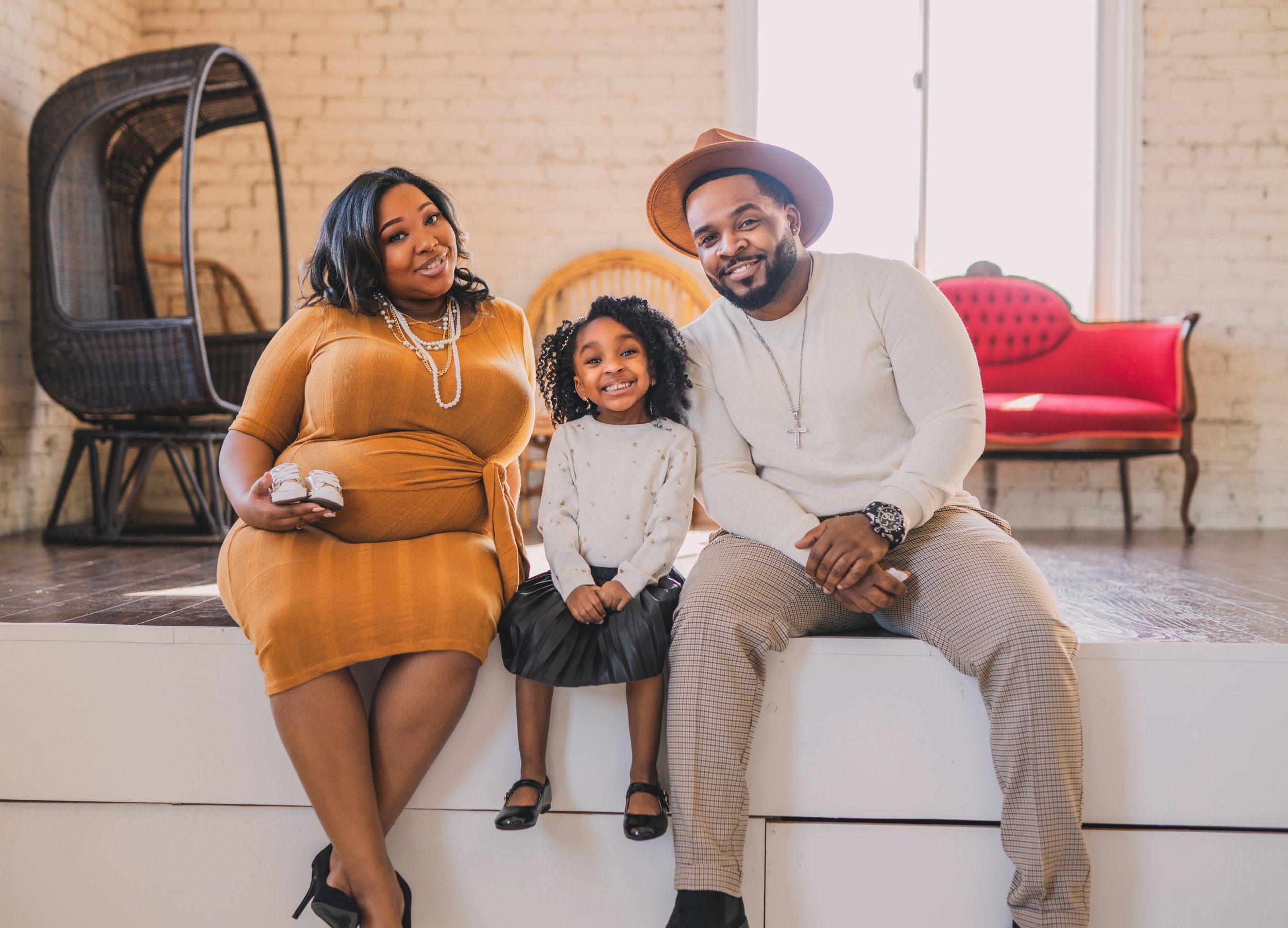 Family of three sitting together indoors, with a woman in a gold dress, a man in checkered pants and hat, and a young girl in a white sweater and black skirt, smiling at the camera.