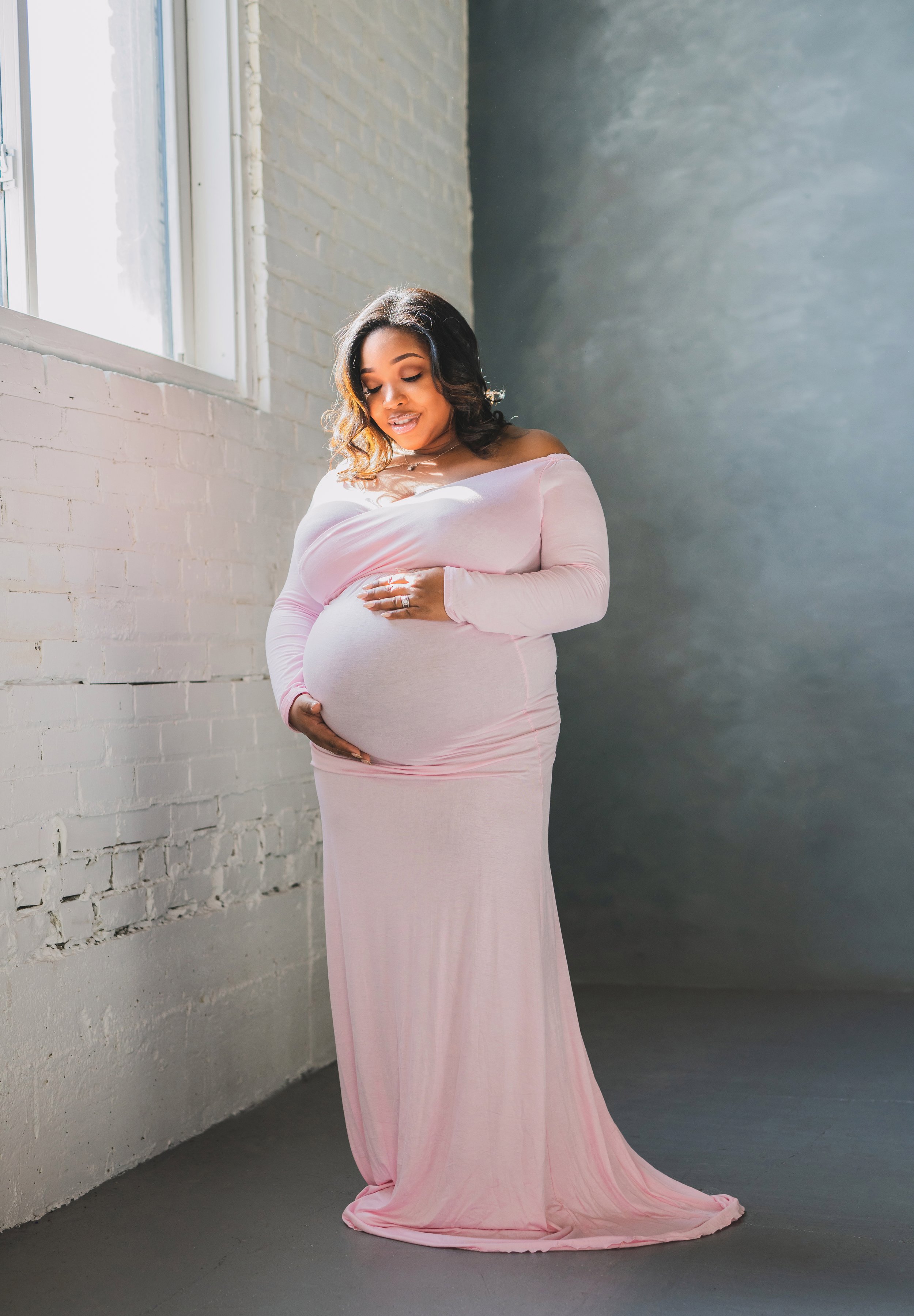 A pregnant woman in a long pink dress standing indoors near a white brick wall and a large window, gently holding her belly and smiling.