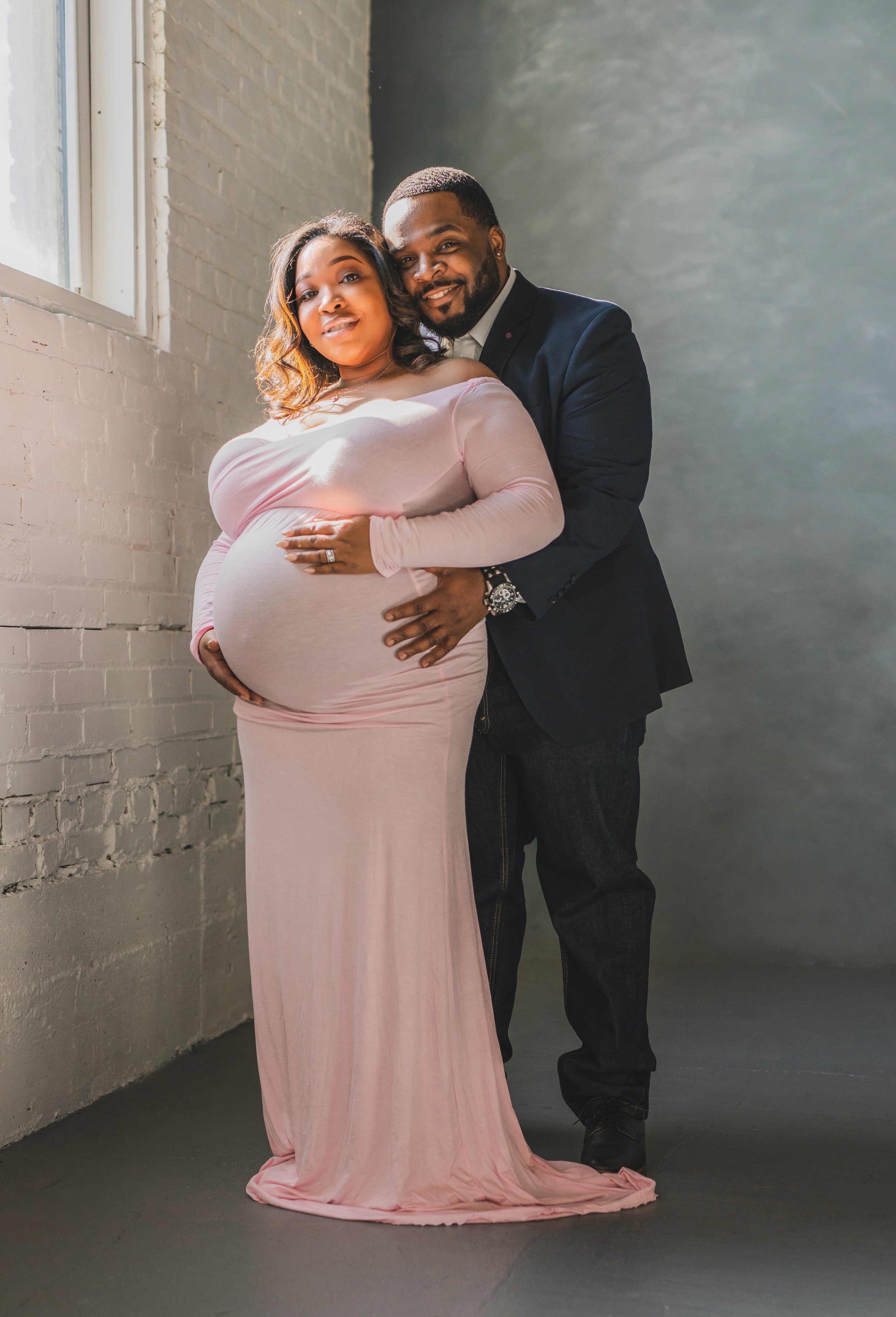 Pregnant woman in a pink dress and a man in a suit standing together inside near a window with a brick wall background.