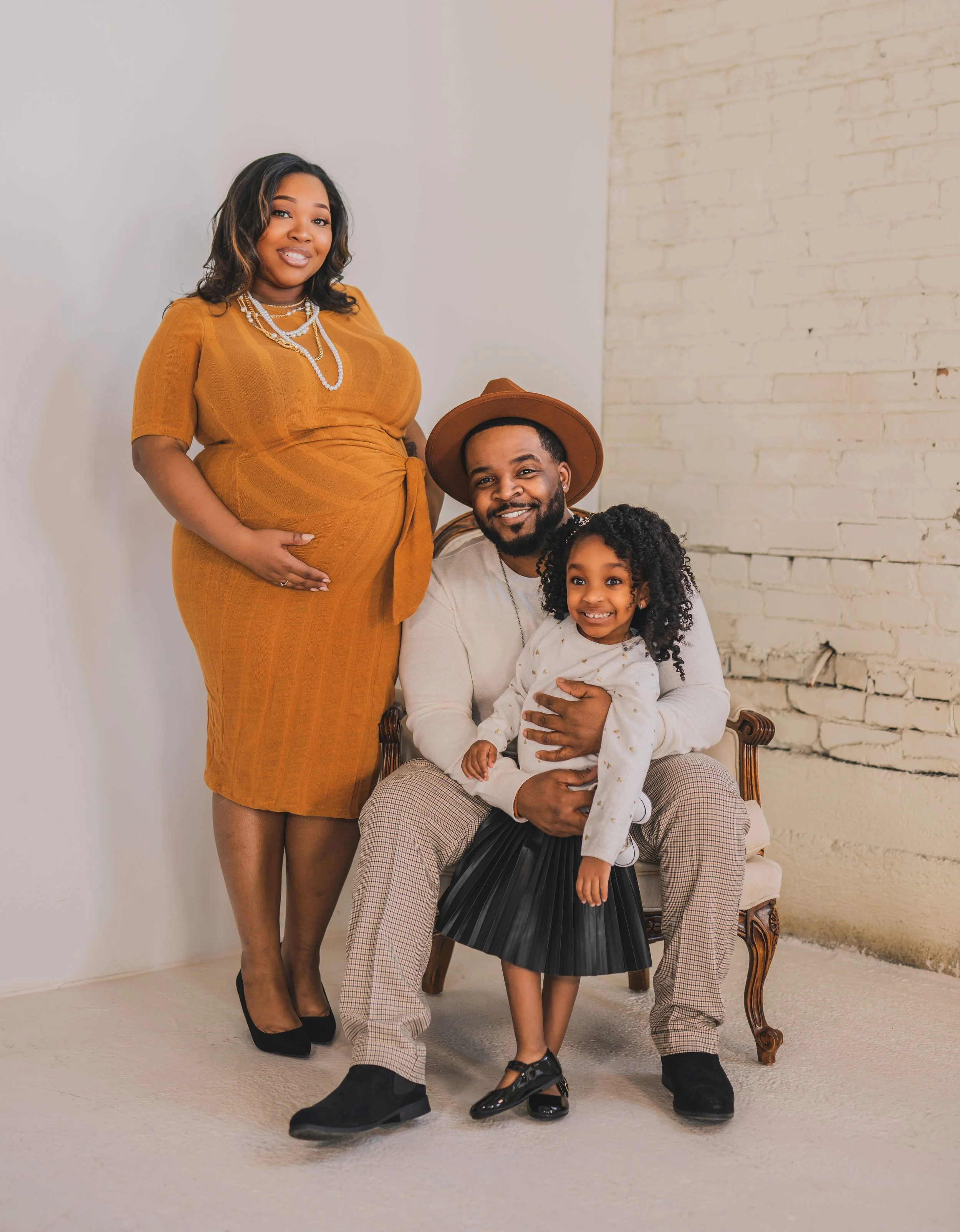 A pregnant woman in a mustard-colored dress standing next to a seated man in a beige hat, a young girl in a white sweater and black skirt, and a white brick wall background.