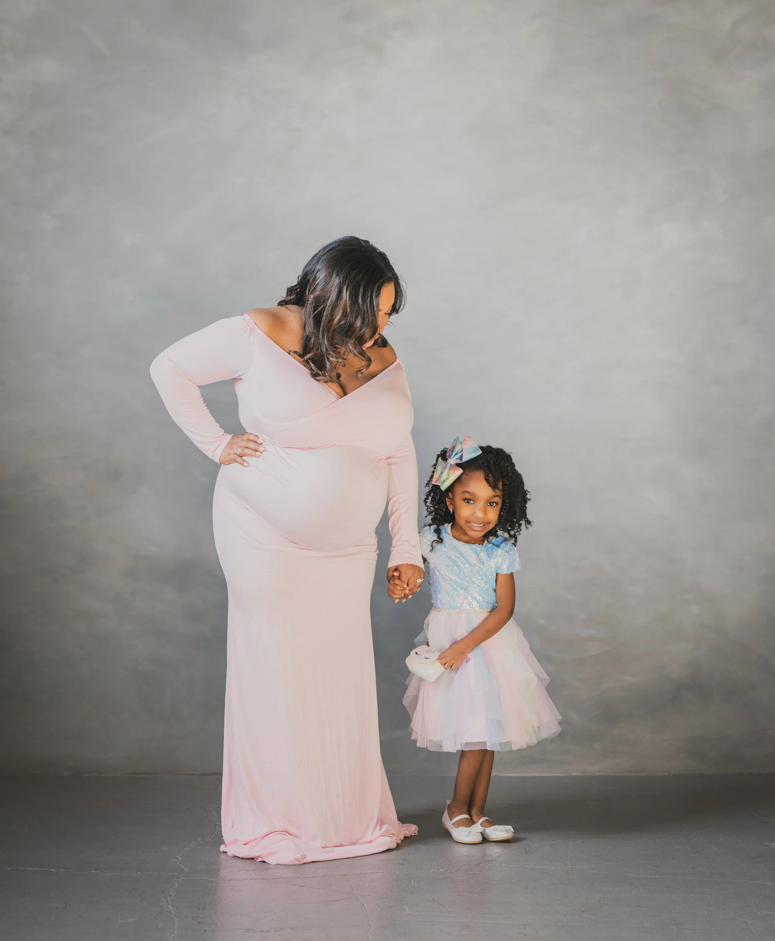 A pregnant woman in a long pink dress holding hands with a young girl in a pastel tulle skirt and a blue top, both standing on a plain gray background.