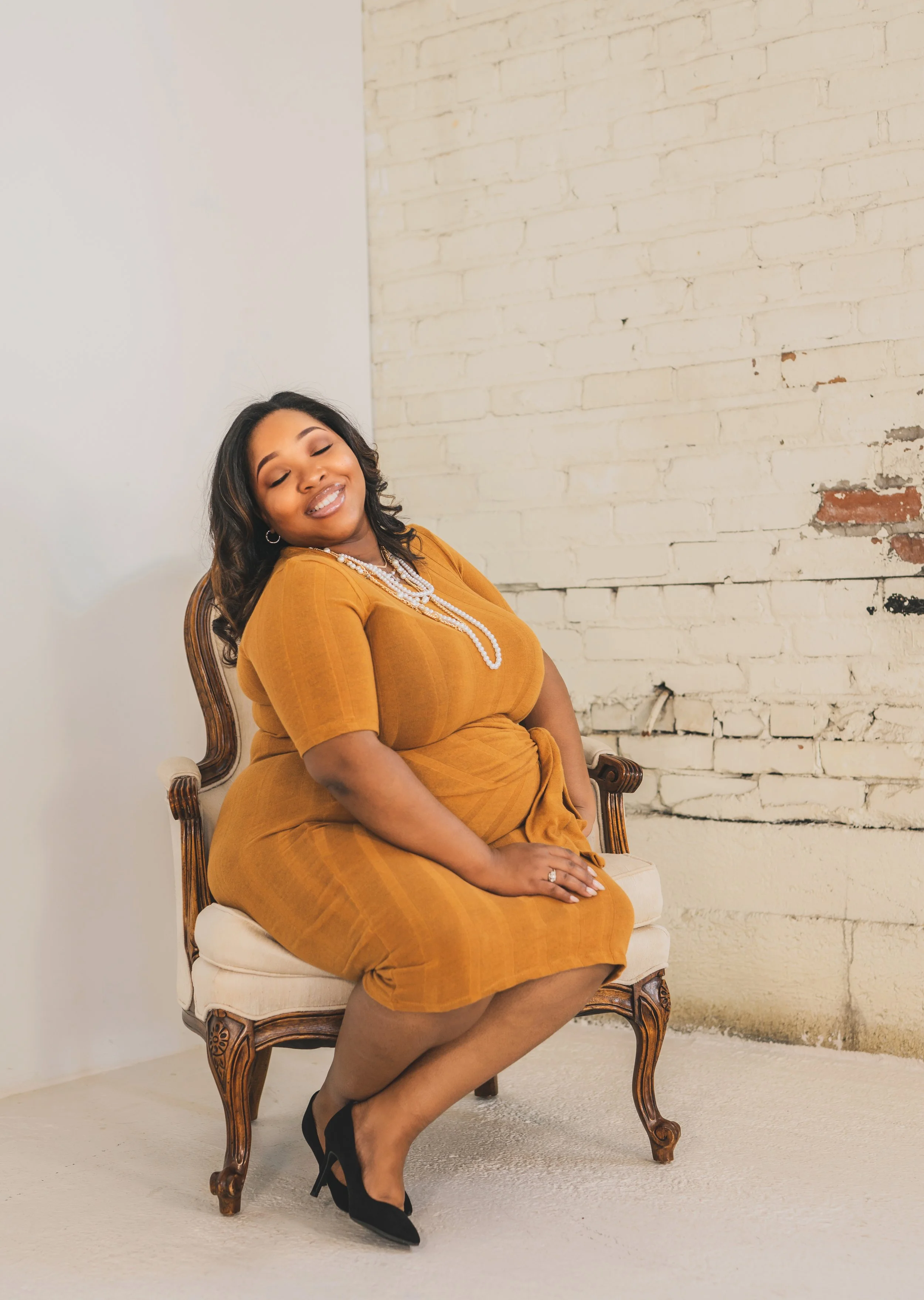 A woman sitting on an elegant wooden chair with a light-colored cushion, wearing a mustard-colored dress and black high heels, posing against a white brick wall with some aged paint and texture, smiling with eyes closed, accessorized with pearl jewel