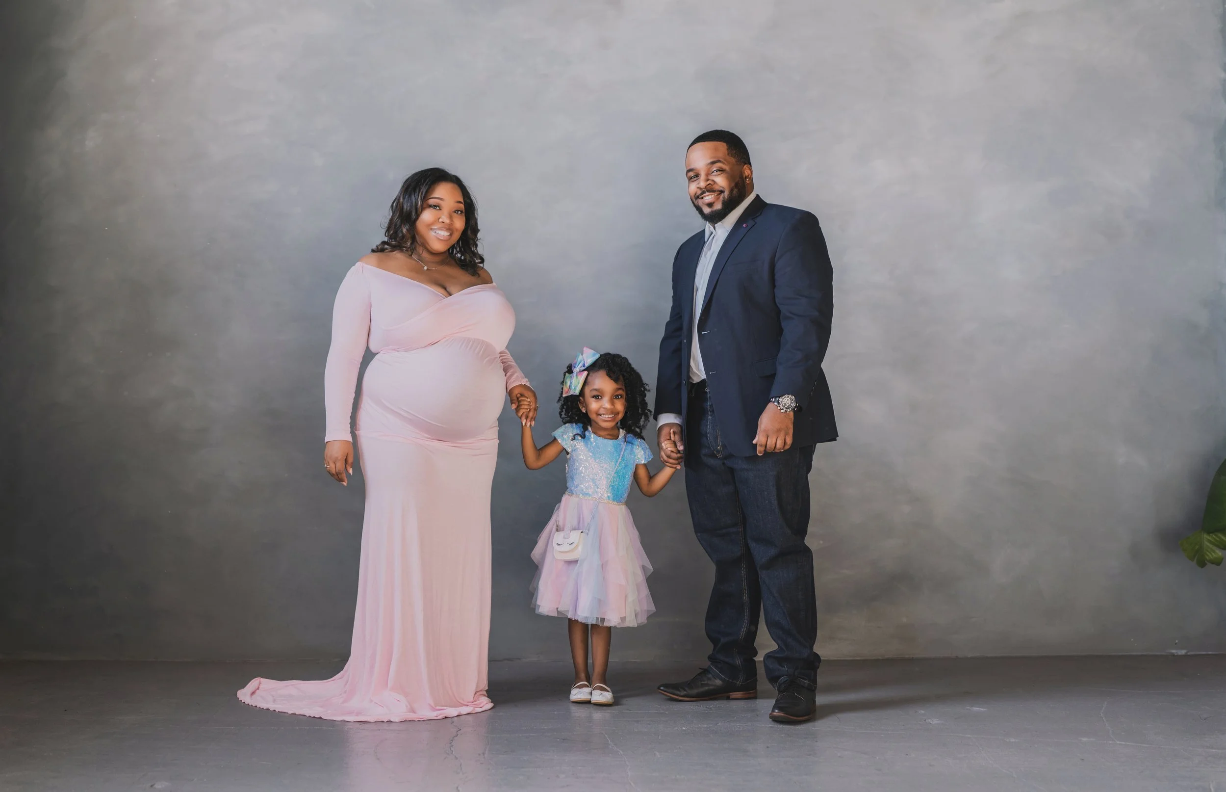 A family of three, a pregnant woman, a man, and a young girl, holding hands and standing together in front of a neutral gray backdrop. The woman is wearing a long pink dress, the girl is wearing a colorful dress with a rainbow bow in her hair, and th