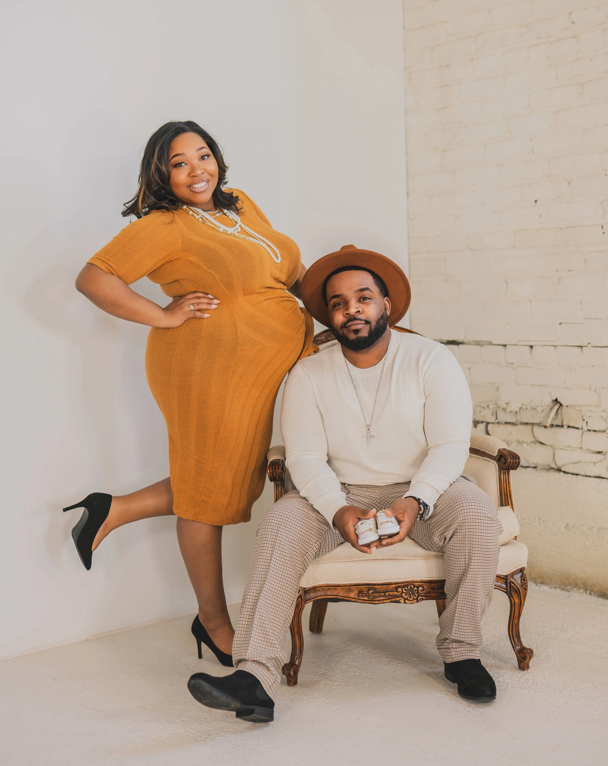 A woman in an orange dress and black heels leans on a man sitting in an antique chair, wearing a cream sweater, plaid pants, and a brown hat, against a white brick wall.