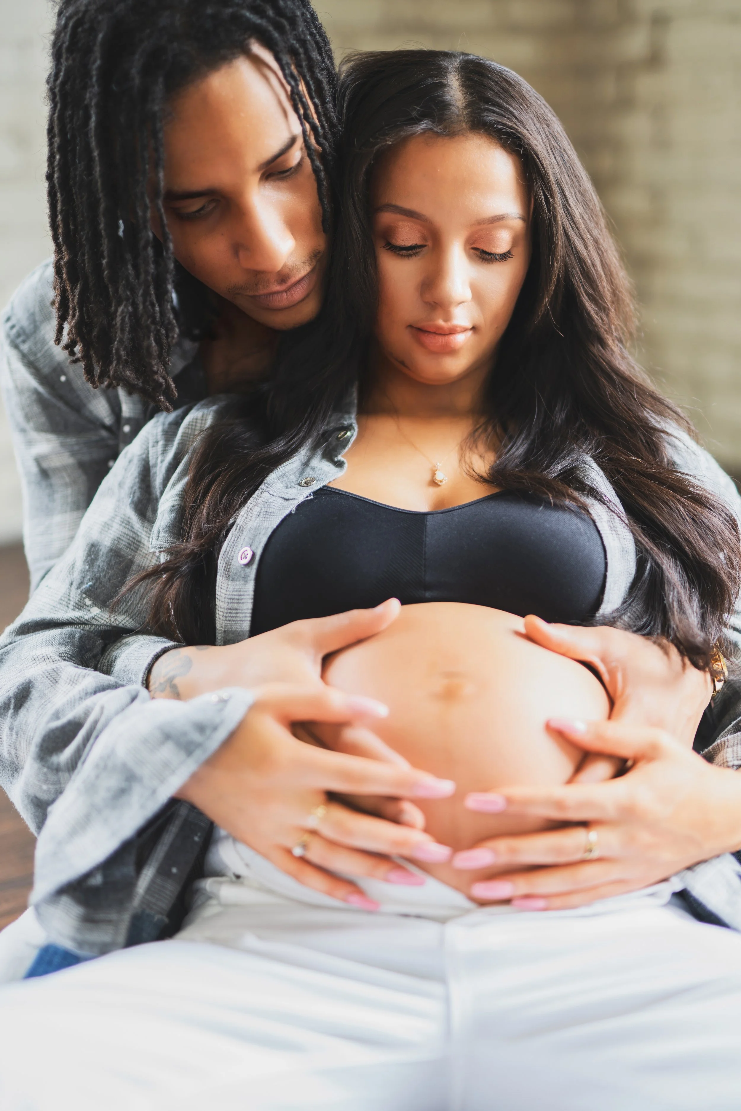 A pregnant woman with long dark hair is sitting with her pregnant belly exposed, her hands resting on her belly. A man with dreadlocks leans over her, embracing her from behind with his hands on her belly, both with calm, gentle expressions.