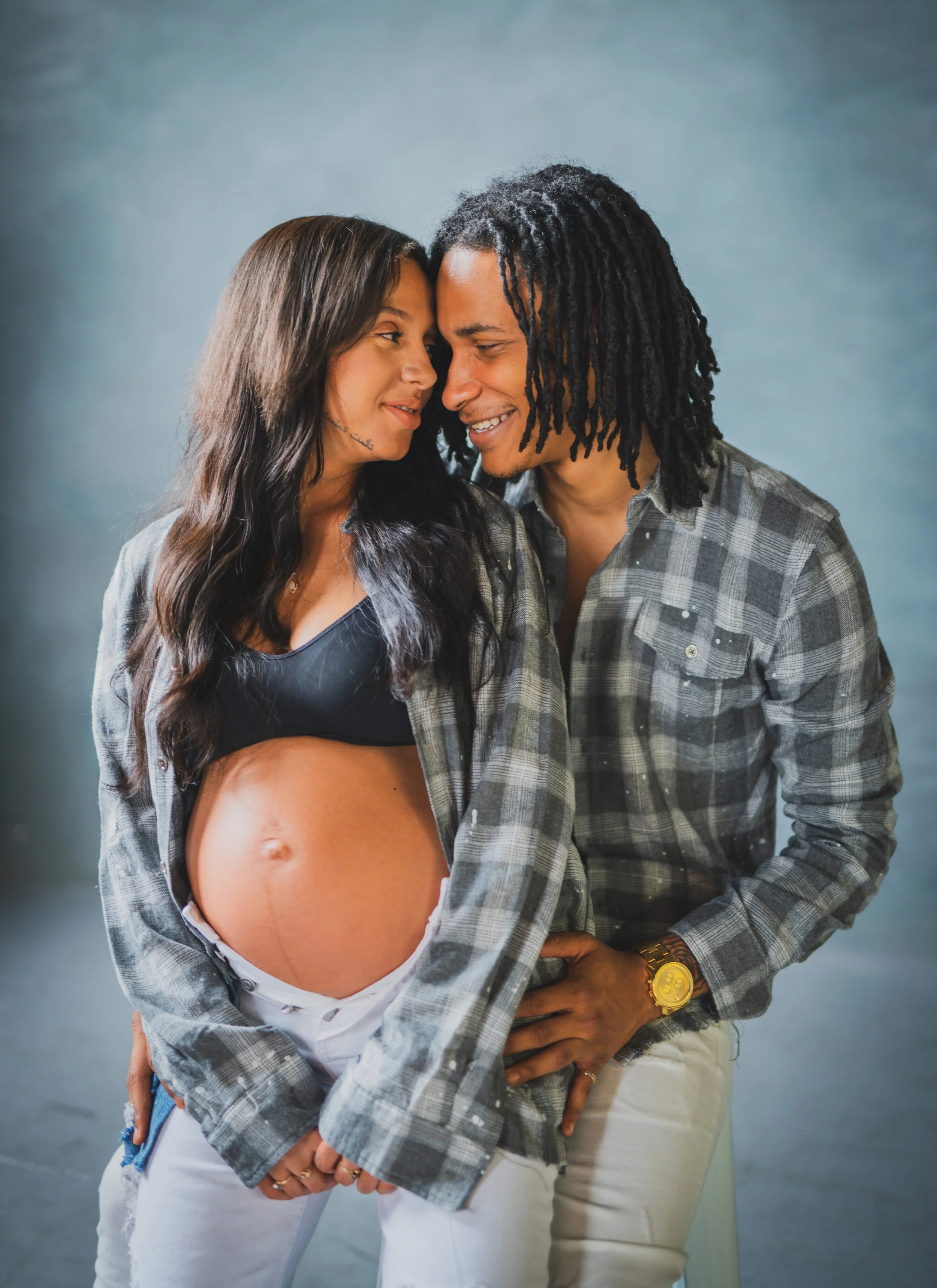 A pregnant woman and her partner softly touching foreheads and smiling, with the woman's pregnant belly visible, wearing casual clothing against a plain background.