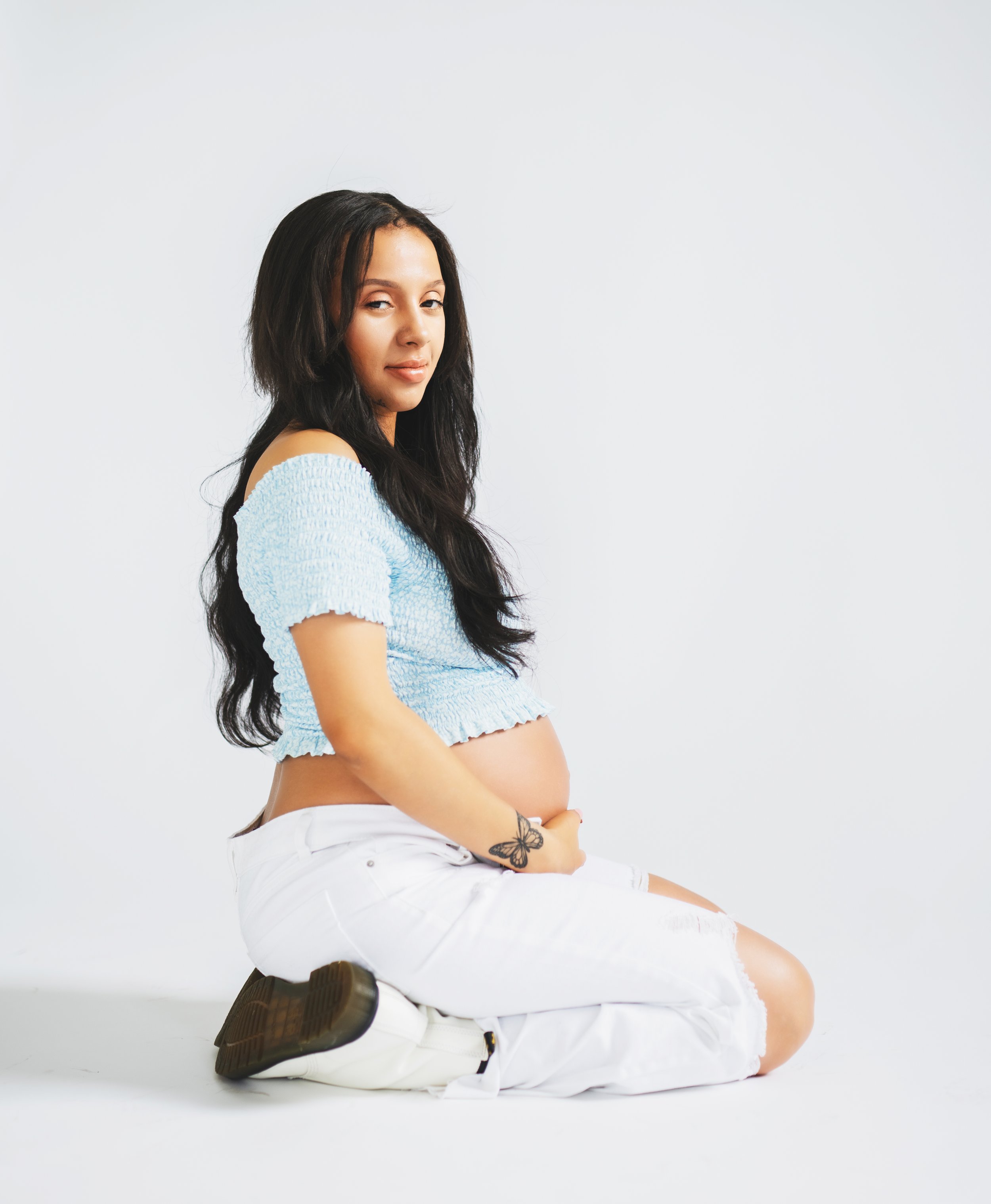 Pregnant woman sitting on the floor, wearing a light blue off-shoulder top and white pants, looking at the camera with a slight smile, against a plain white background.