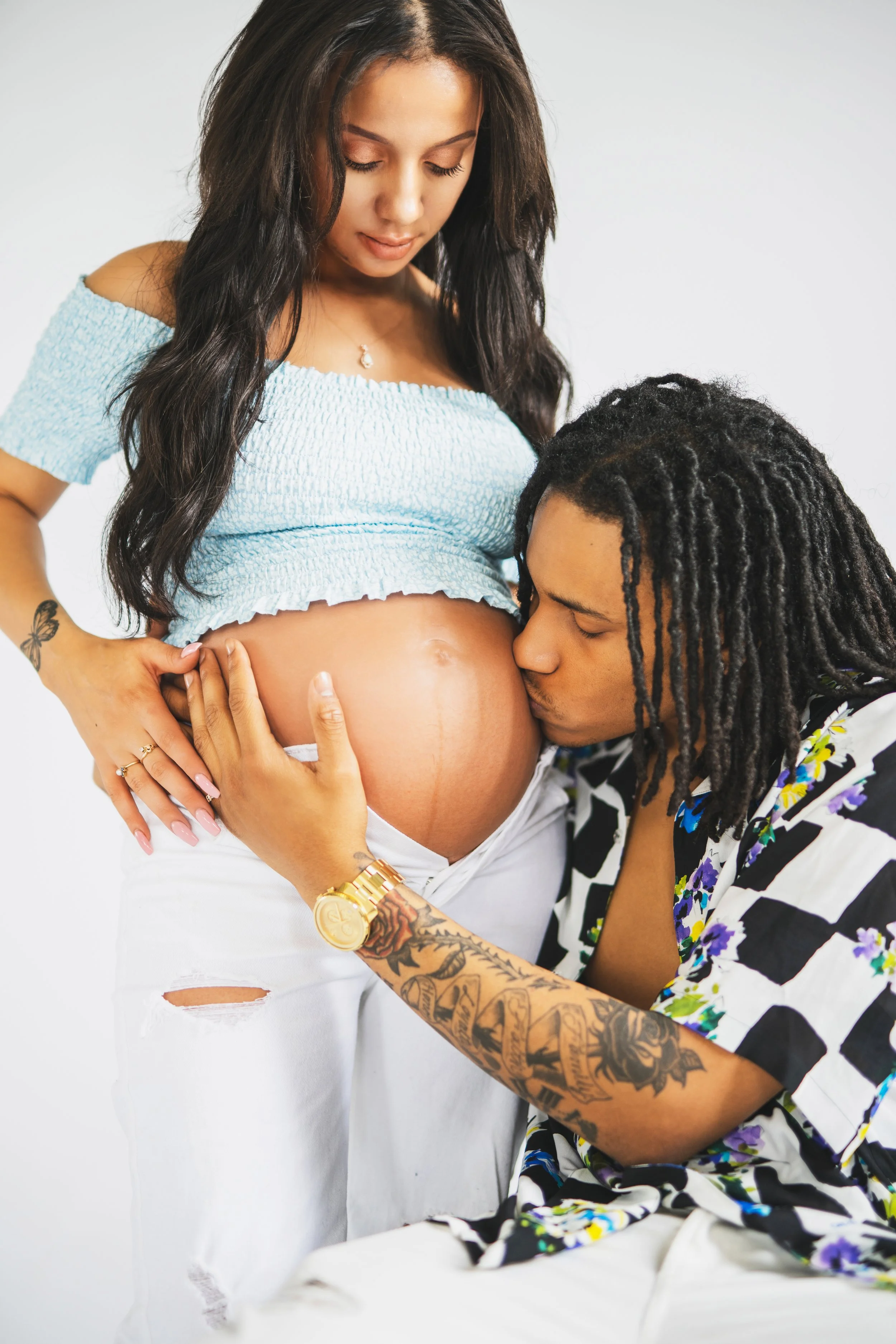 A pregnant woman with long dark hair wearing an off-shoulder blue top and white pants with a tear, standing while a man with dreadlocks, tattoos, and a colorful shirt kisses her baby bump. She is gently holding her belly and looking down at it, while