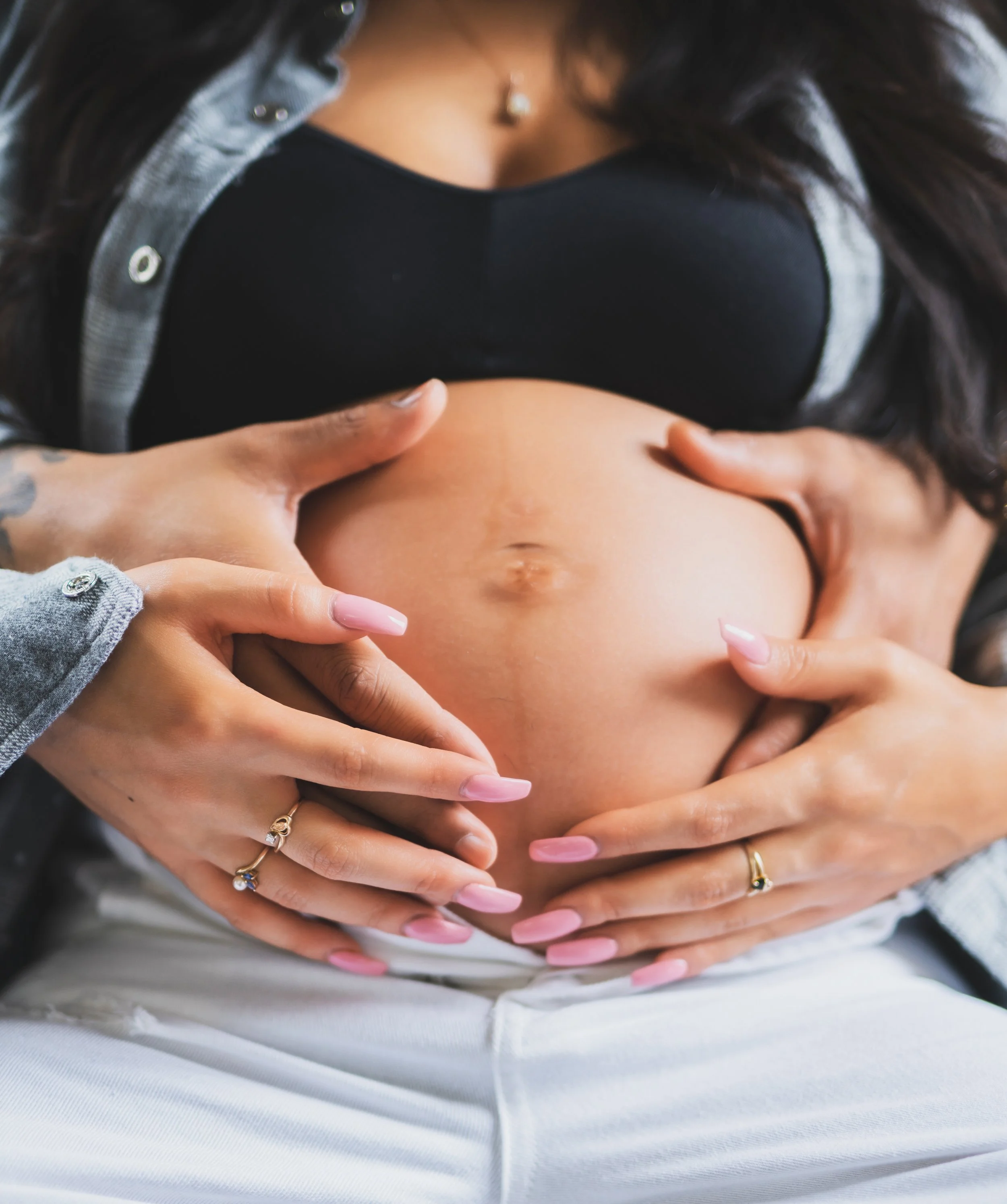 Close-up of a pregnant woman's belly being cradled by two hands with jewelry, including rings and nail polish.