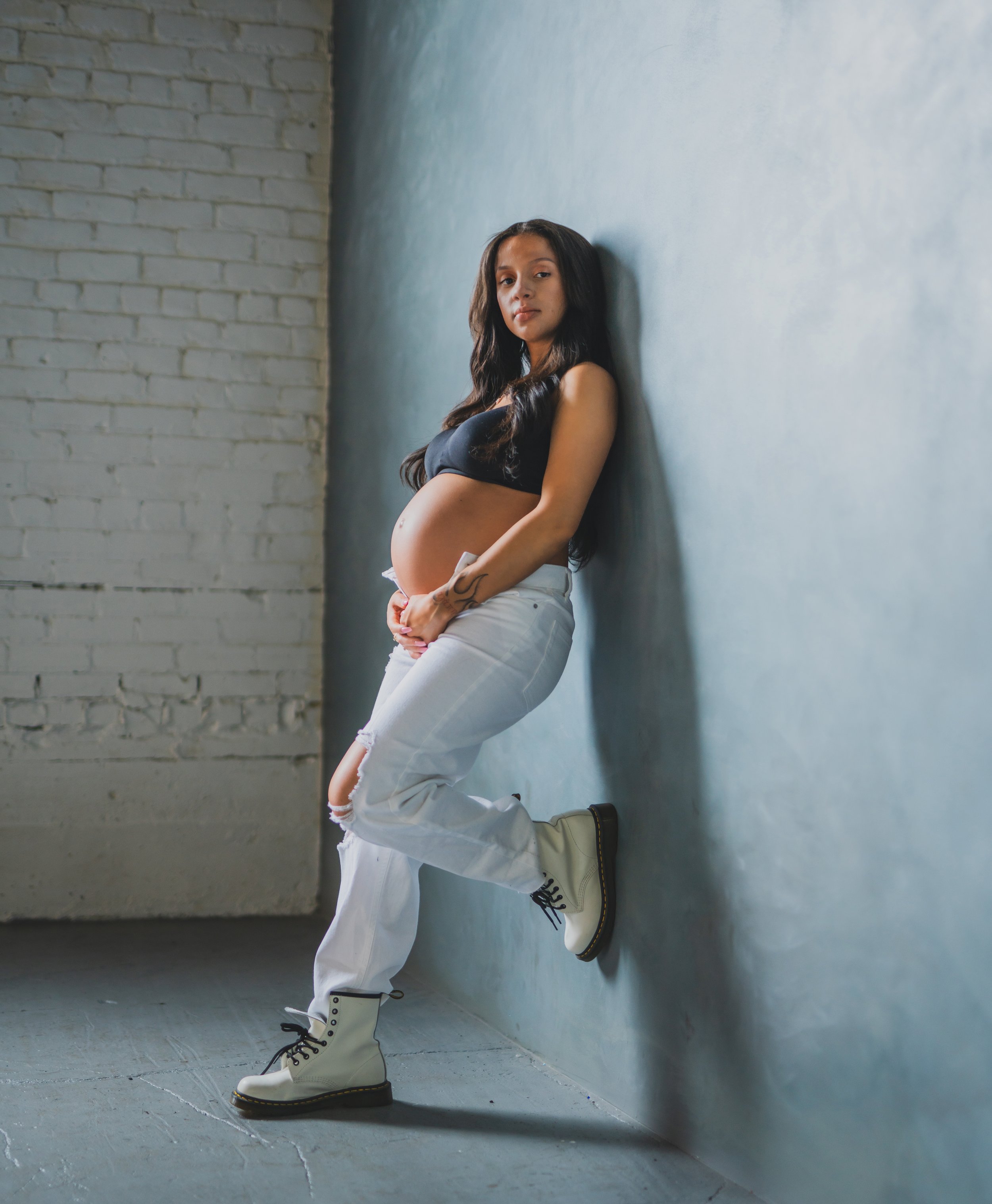 Young pregnant woman leaning against a wall with hands on her belly, wearing a black top, ripped white jeans, and white boots.