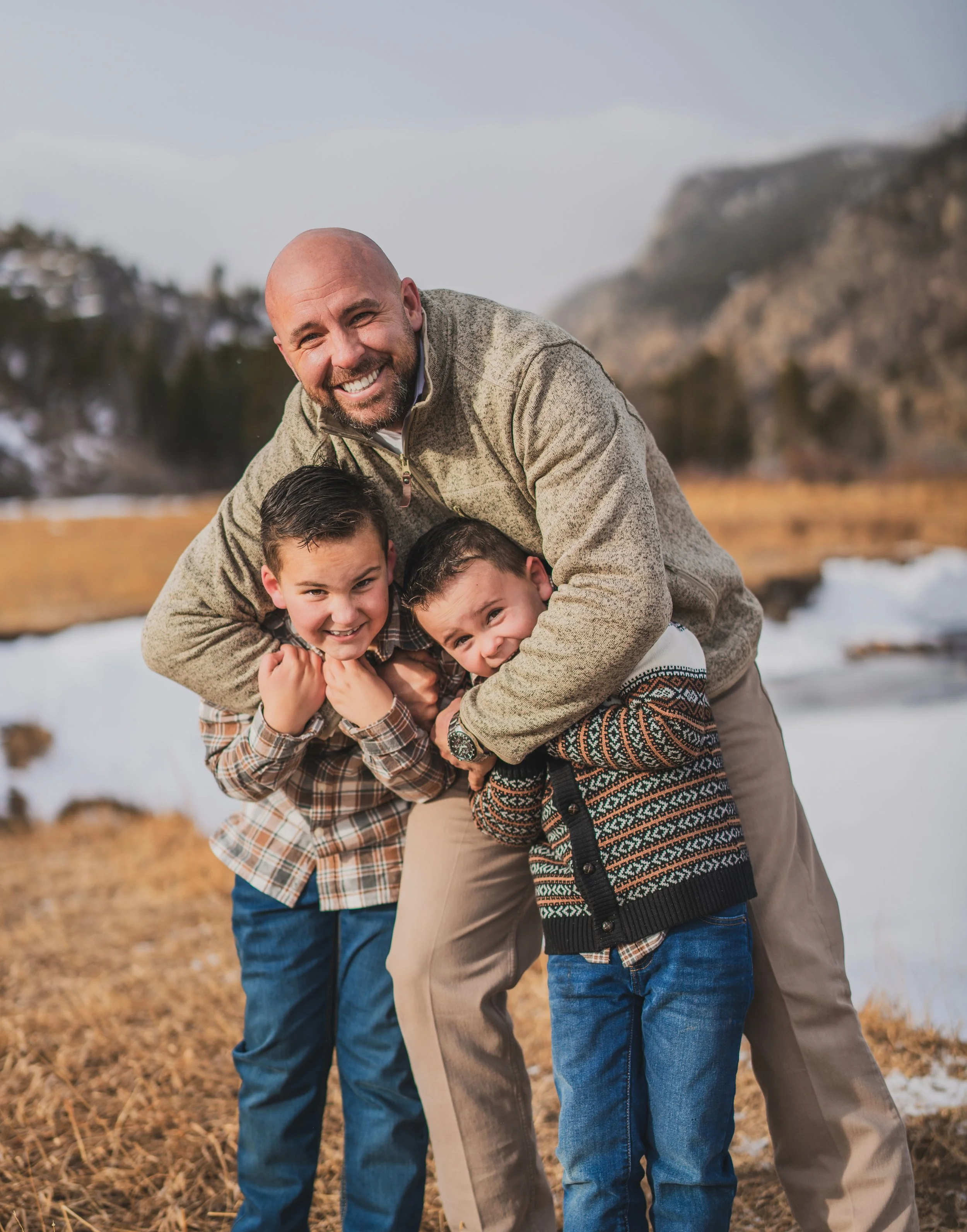 A smiling man hugging two young boys outdoors in a field with mountains in the background, during winter.