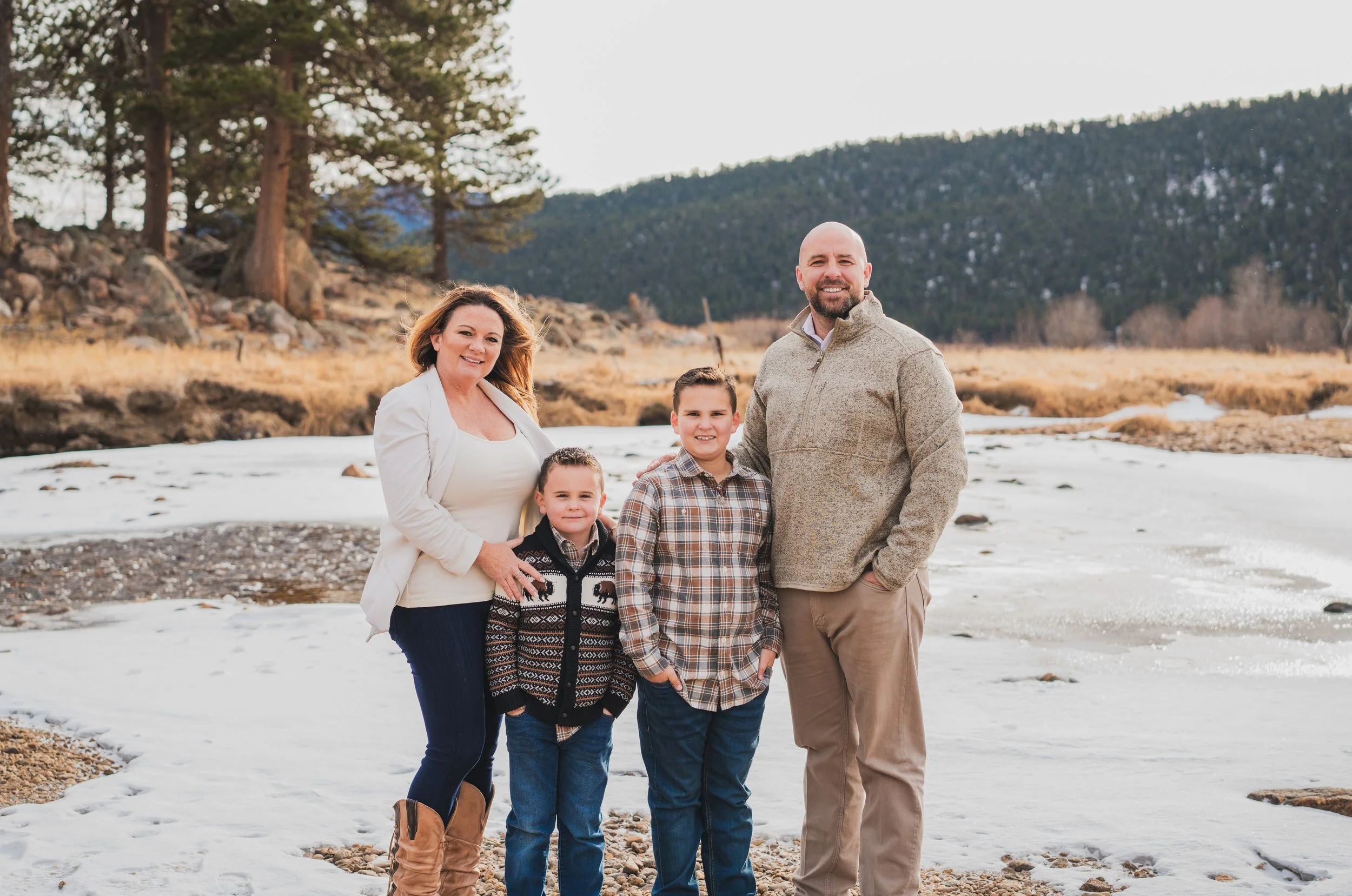 Family of four standing on a snowy riverbank with trees and mountains in the background