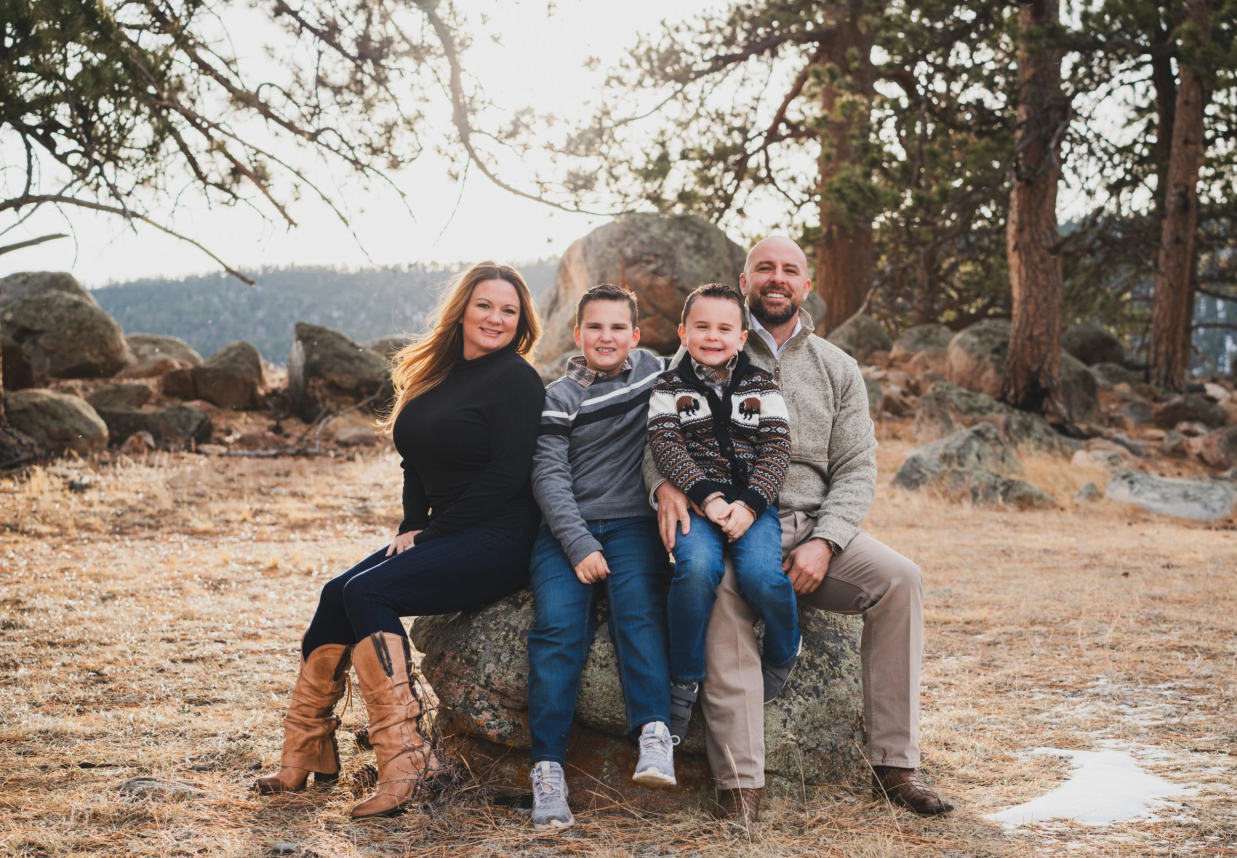 Family of four sitting on a rock outdoors in a forested area with trees and rocks in the background.