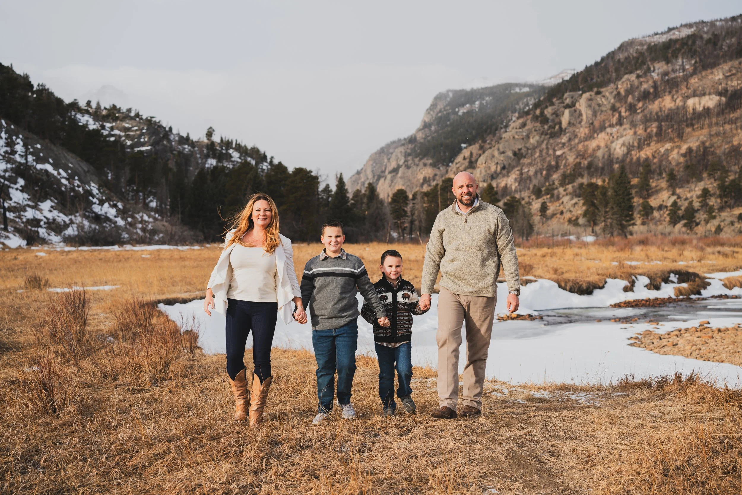 Group of two adults and two children walking hand-in-hand outdoors in a mountainous area with a partly frozen stream, dry grass, trees, and snow-covered hills in the background.