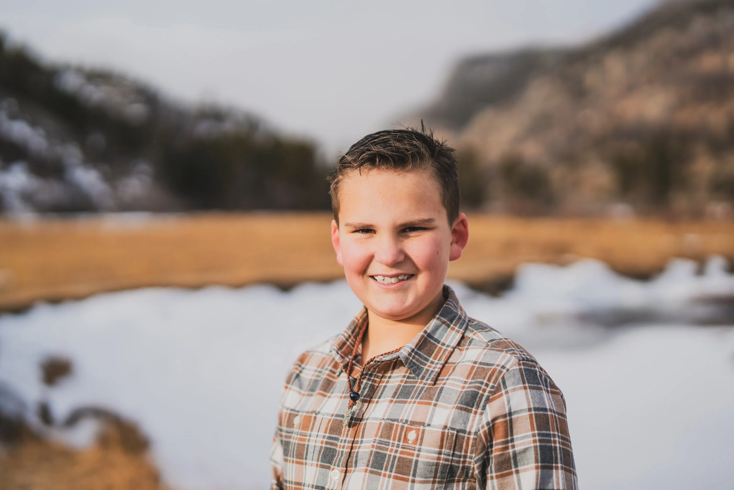 Young boy smiling outdoors in a snowy, mountainous landscape, wearing a plaid shirt.