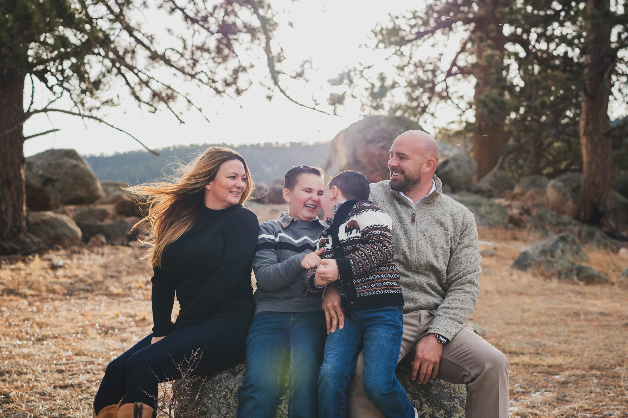 Family of four outdoors sitting on a rock in a natural landscape, smiling and laughing together in autumn.