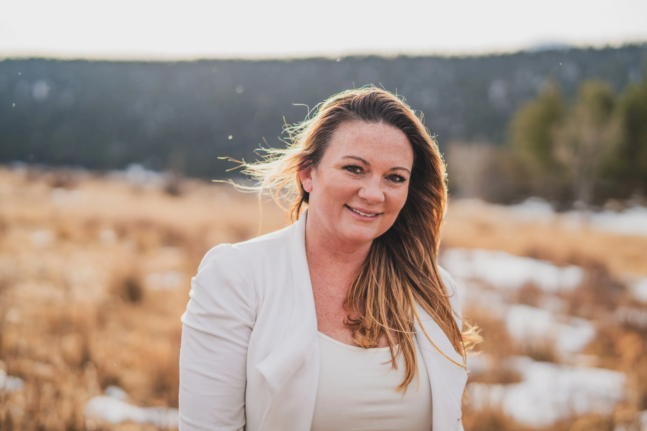 A woman with long brown hair, wearing a white top and jacket, smiling outdoors in a field with patches of snow and a forested hill in the background.