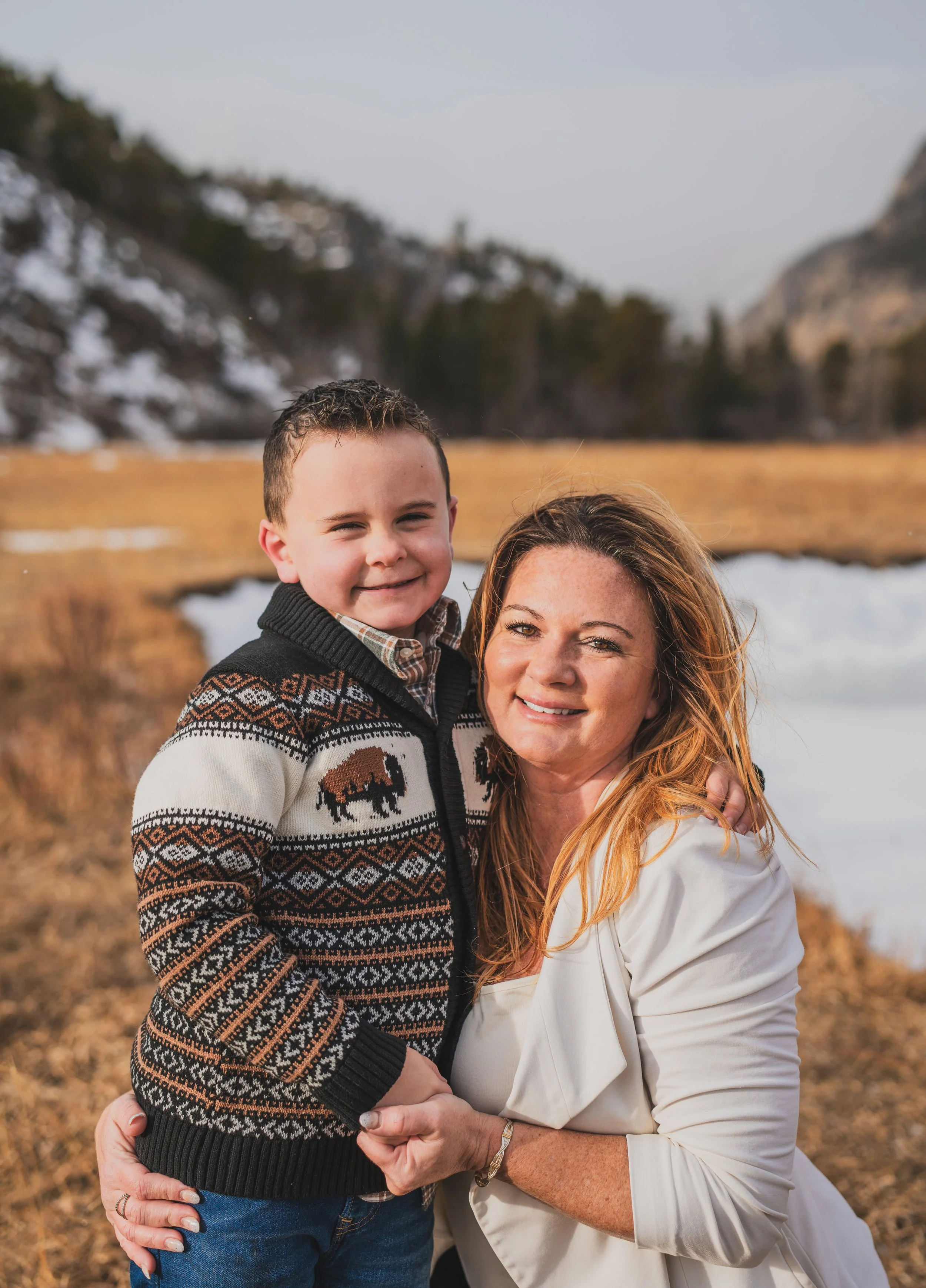 A woman with red hair and a young boy with dark hair standing outdoors near a river or lake, with mountains and trees in the background. The woman is smiling and the boy is smiling, both dressed warmly.