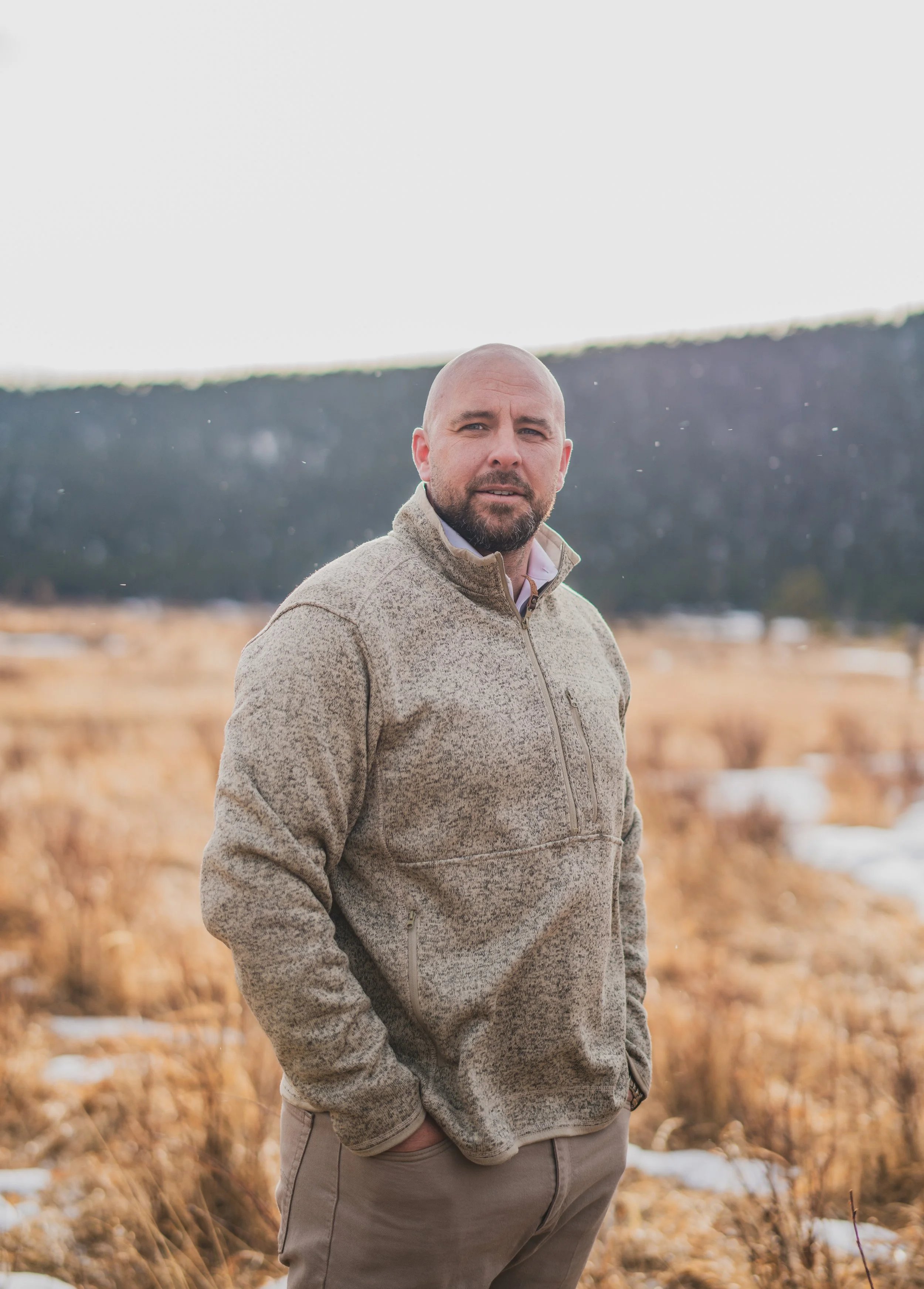 A man with a bald head and beard standing outdoors in a field with dry grass and snow patches, wearing a beige fleece jacket and gray pants, with a mountain and cloudy sky in the background.