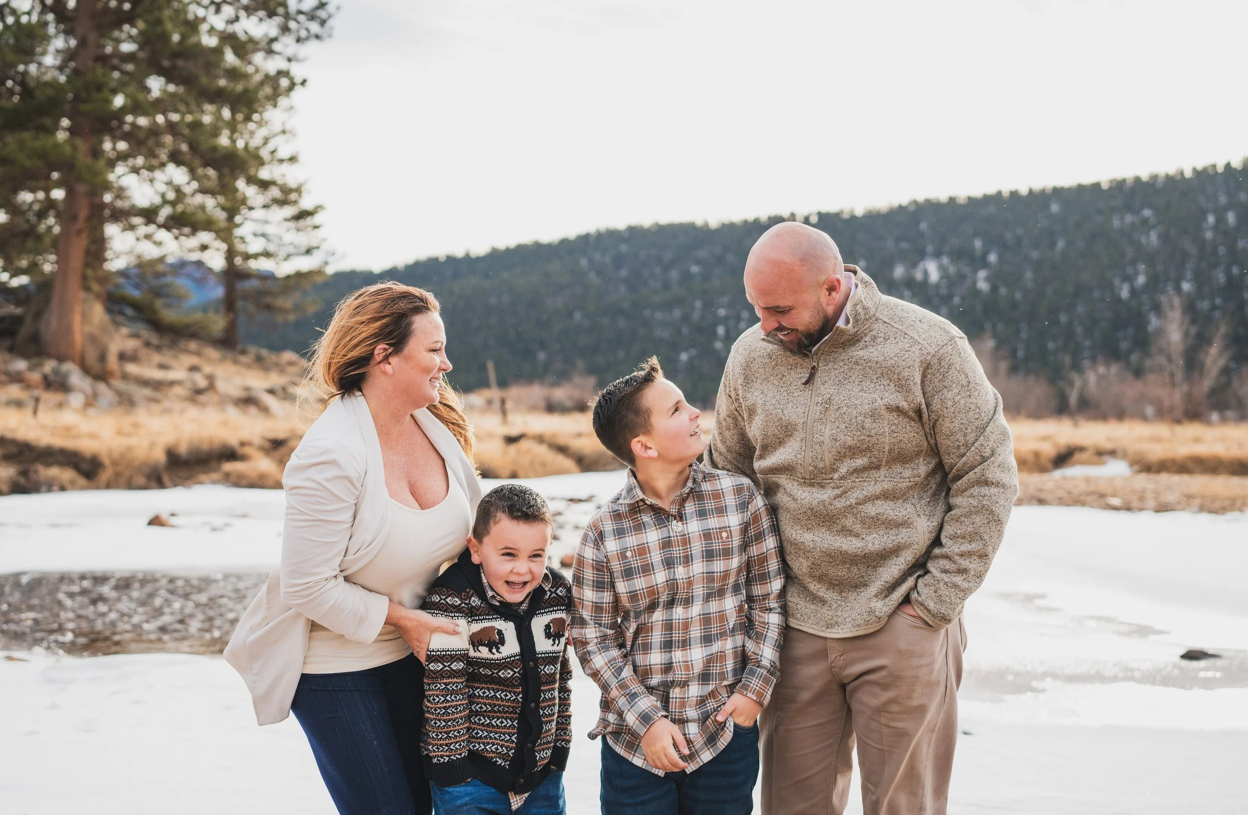 A family of five, including two children, stands outdoors on a snowy landscape with mountains in the background. They are smiling and enjoying each other's company.