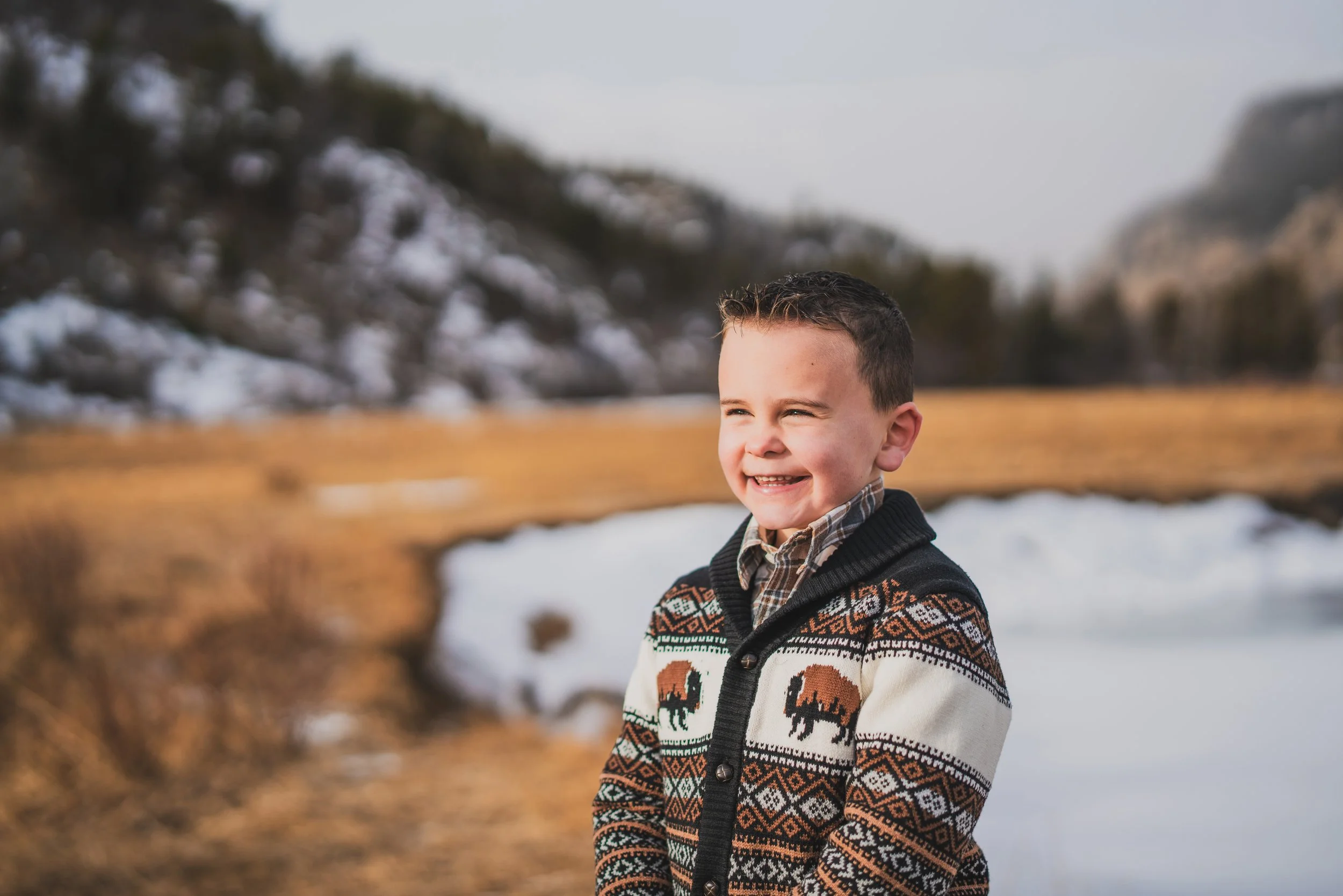 A smiling young boy standing outdoors in a winter landscape with snow and trees in the background, wearing a patterned sweater with bison designs.